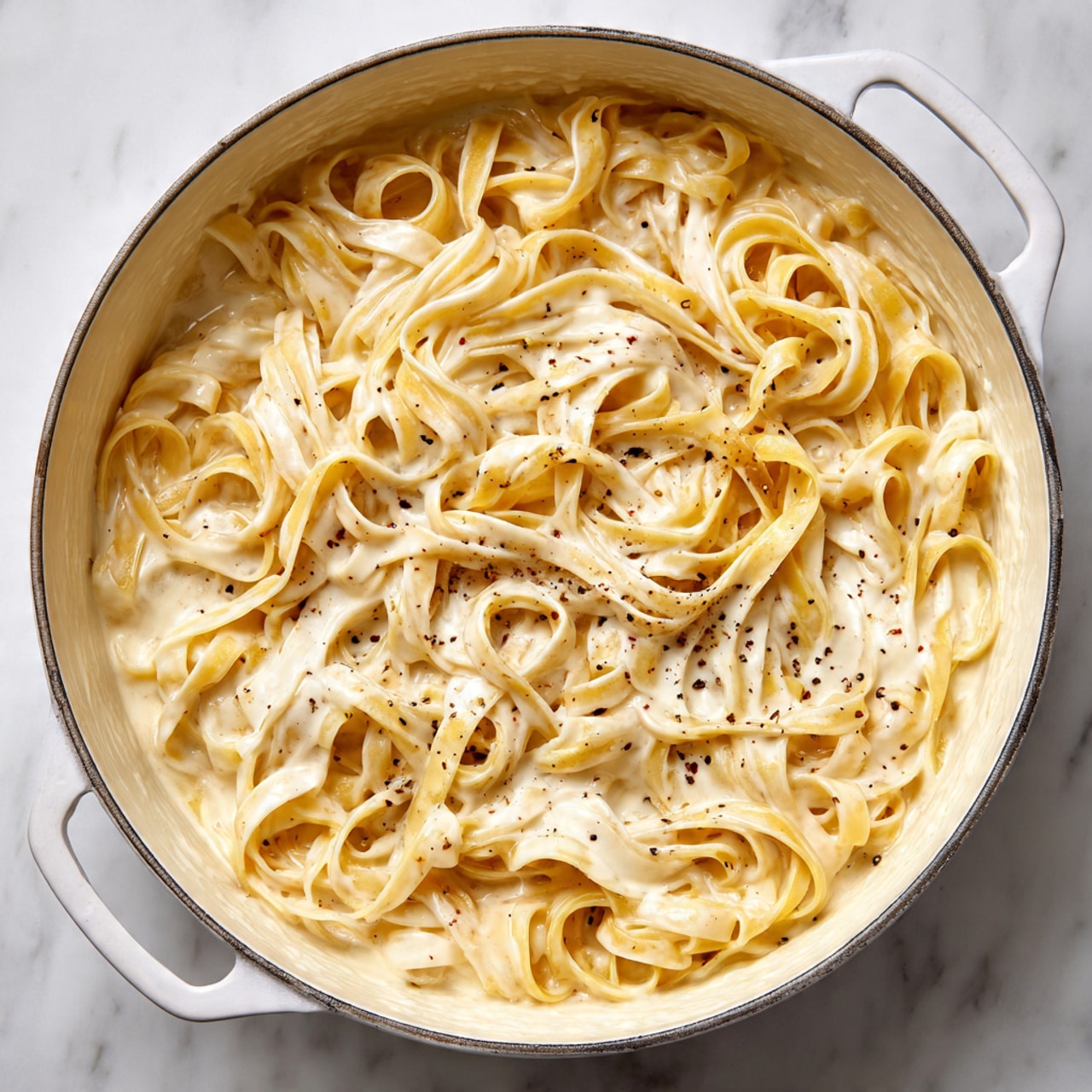 The image shows a large white pot filled with creamy fettuccine pasta. There is one main layer of creamy white sauce coating long, flat pasta strands that are thick and slightly curled in several places. The creamy sauce has small dark specks of seasoning throughout, creating a rich texture. The pot sits on a white marbled surface, and the edges of the pot are visible, showing the white interior. The sauce looks smooth and evenly mixed with the pasta, with some pasta strands piled on top of others in a loose, natural swirl. Photo taken with an iphone --ar 4:5 --v 7