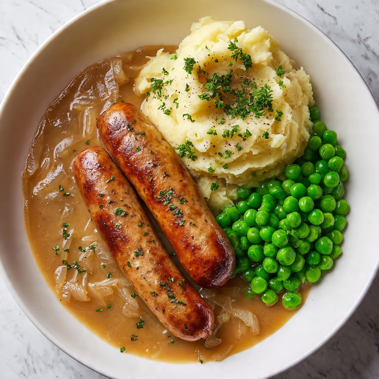 A white bowl contains the dish with three main layers: at the bottom is a smooth, light brown onion gravy with visible small chunks of onions evenly spread across the base; on top of the gravy, there are two browned sausages placed diagonally, with a shiny and crispy skin displaying herbs sprinkled on top; next to the sausages on one side, there is a mound of creamy, pale yellow mashed potatoes topped with parsley and black pepper, and on the other side, a cluster of bright green peas fills the space, some overlapping the mashed potatoes, all placed on a white marbled textured surface; the image is bright and clear, photo taken with an iphone --ar 4:5 --v 7