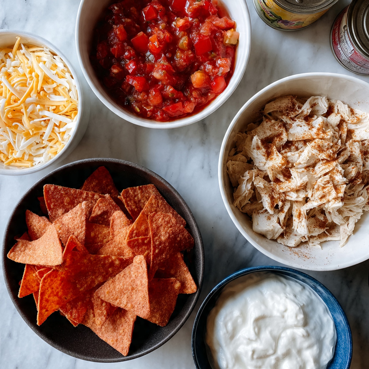 The image shows several bowls arranged on a white marbled surface with two cans of cream of chicken soup in the background. In the upper left, there is a white bowl filled with shredded mixed yellow and white cheese. Next to it on the right, a white bowl holds chunky red salsa with visible pieces of tomatoes and other ingredients. Below, a large black bowl is filled with broken red tortilla chips. To the right, a white bowl contains shredded light-colored chicken topped with brown seasoning. In the front right, a blue bowl contains thick white sour cream with some texture visible. The overall scene displays raw ingredients for a layered dish. Photo taken with an iphone --ar 4:5 --v 7