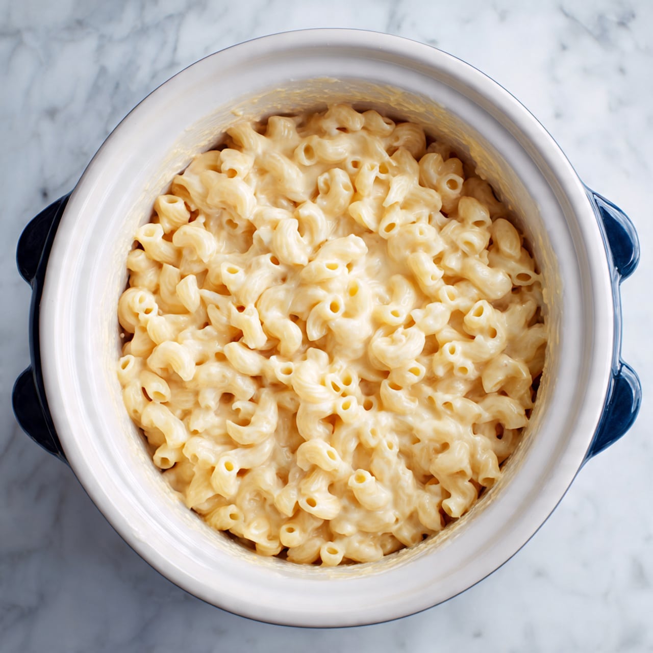 A white round slow cooker filled with creamy, light yellow macaroni pasta. The macaroni is elbow-shaped and fully coated in a smooth cheese sauce that has a slightly shiny, soft texture. The sauce looks thick and rich, covering all the pasta pieces evenly. The edge of the slow cooker shows a bit of sauce residue. The slow cooker has dark blue handles on both sides and is placed on a white marbled surface. photo taken with an iphone --ar 4:5 --v 7