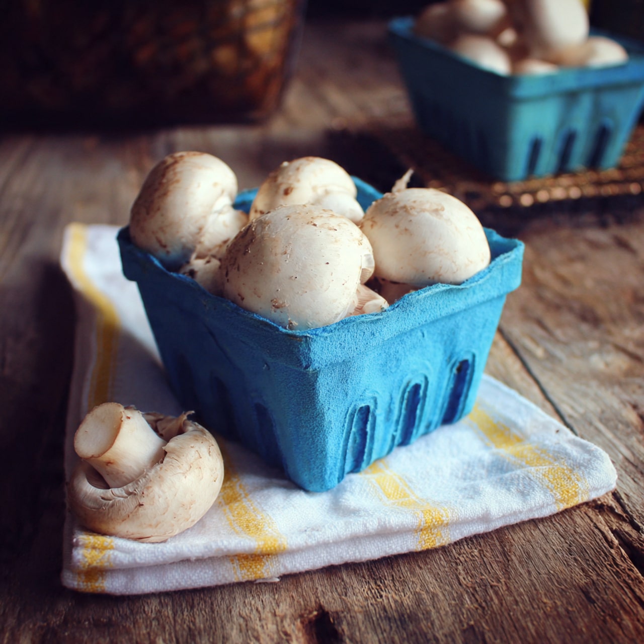 The image shows a small group of fresh white mushrooms inside a blue plastic container placed on a rustic wooden surface. One mushroom rests partly on a white and yellow striped cloth, positioned in the lower right part of the frame. The background is softly out of focus, including a second container of mushrooms in the top right corner. photo taken with an iphone --ar 4:5 --v 7
