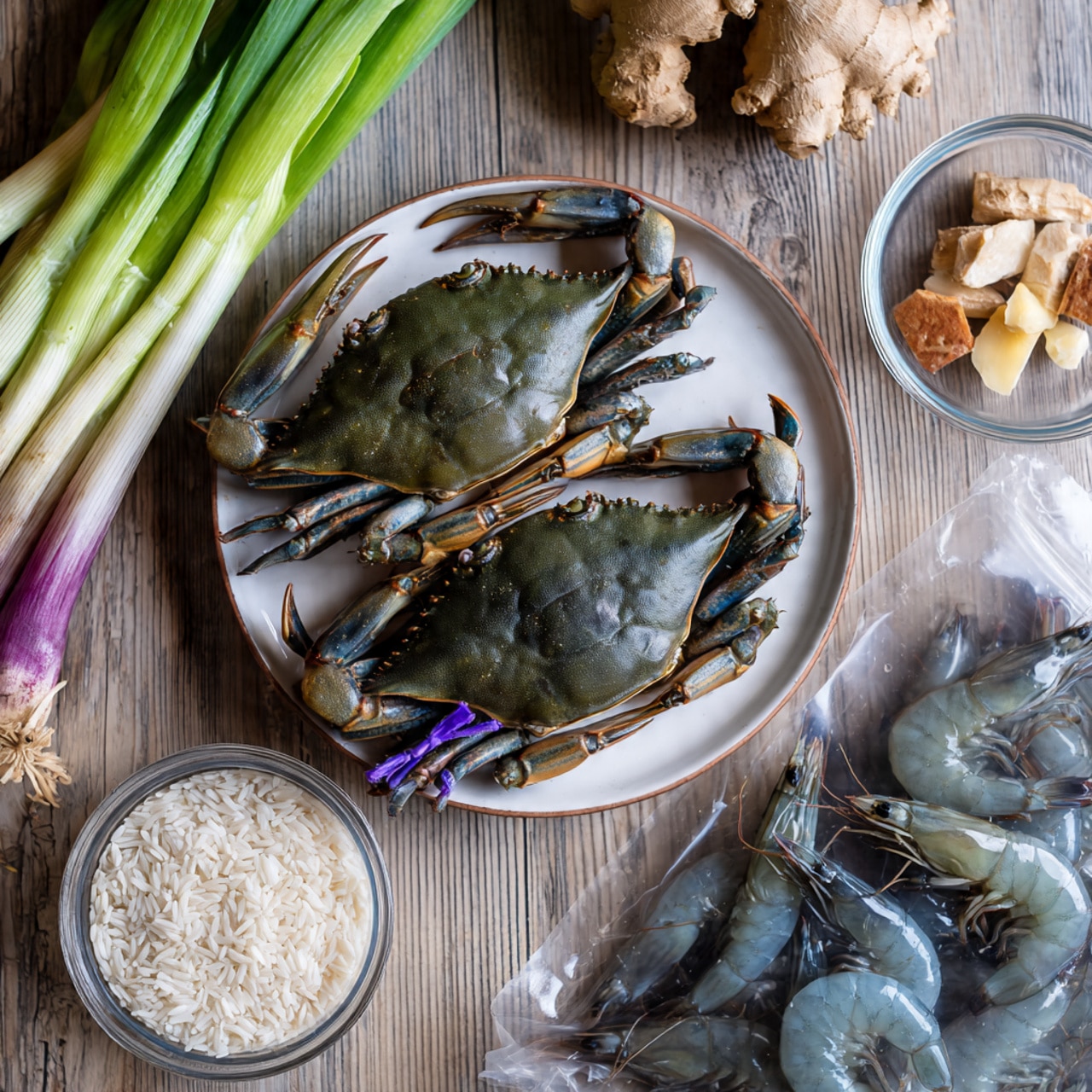 Two dark green live crabs with tied purple bands on their claws sit on a white plate, placed on a wooden textured surface. To the right, a clear glass bowl holds small, light beige dried seafood pieces and a small piece of fresh ginger. Below it, a small round transparent bowl is filled with white rice grains. Fresh green onions with white and purple bases and green stems, along with celery stalks, lie diagonally on the left side. At the bottom right, a plastic bag contains several raw dark gray prawns. photo taken with an iphone --ar 4:5 --v 7