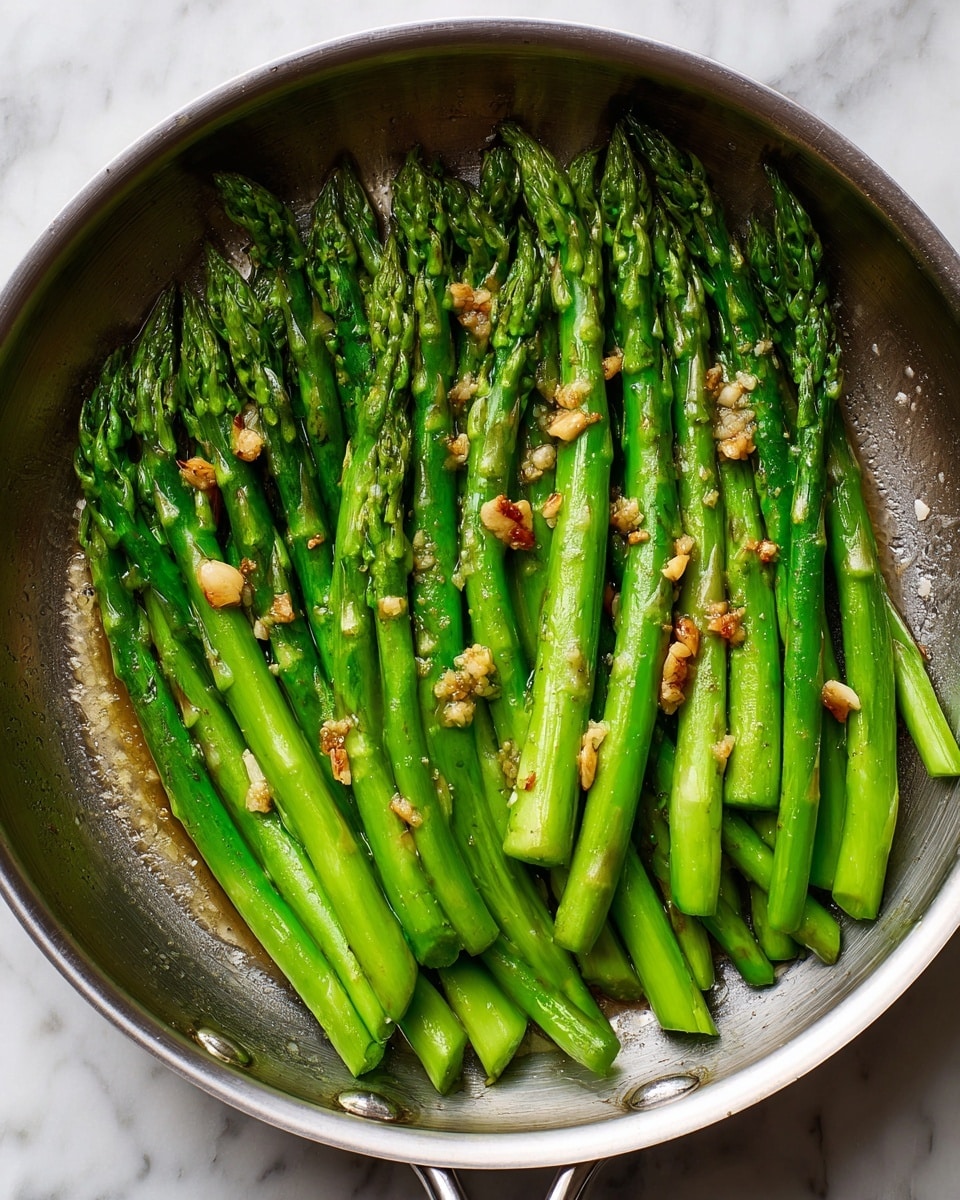 A silver frying pan filled with cooked green asparagus stalks neatly aligned side by side, with small bits of light brown garlic scattered on top. The asparagus shows a fresh, vibrant green color with slightly darker tips and a shiny, slightly oily surface. The pan sits on a white marbled surface, creating a clean and fresh look. Photo taken with an iphone --ar 4:5 --v 7