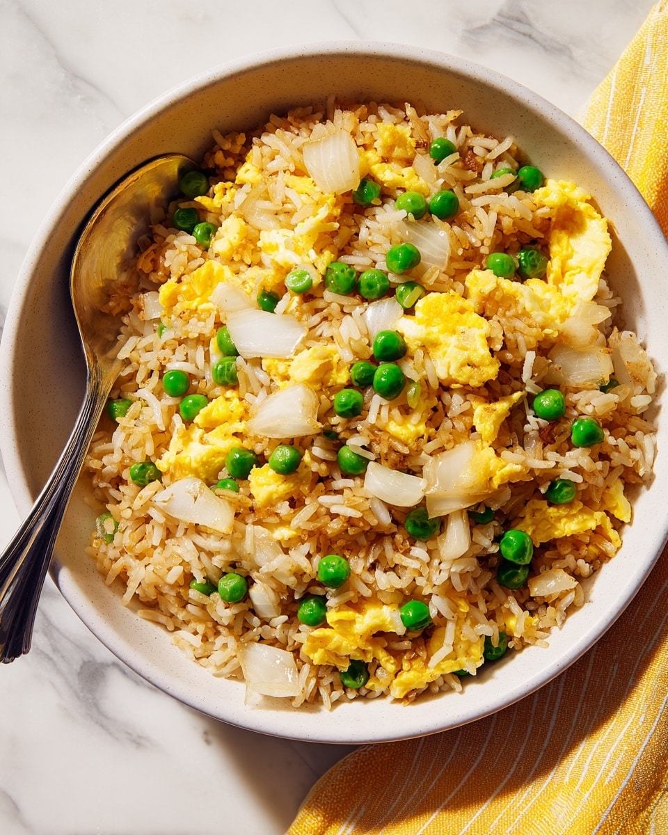 A bowl of fried rice with three main layers visible: the base layer is made of light brown fried rice grains mixed closely with small pieces of scrambled egg in a soft yellow shade, scattered evenly throughout. The middle layer consists of medium-sized chunks of translucent white cooked onions, creating a slight contrast in texture and color. The top layer has bright green peas distributed evenly across the dish, adding pops of vibrant color. The food is served in a white bowl with a smooth, slightly speckled texture, and a silver spoon rests inside on the left side of the bowl. The background is a white marbled surface with a yellow striped cloth underneath the bowl. Photo taken with an iphone --ar 4:5 --v 7
