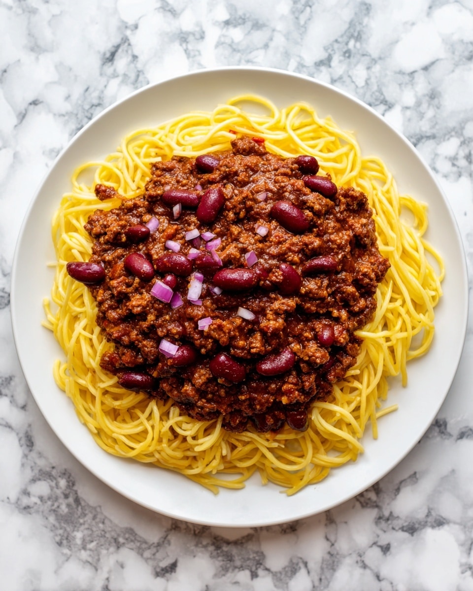 A white plate holds a base layer of yellow cooked spaghetti noodles, slightly twisted and spread out. On top of the noodles is a thick layer of dark brown chili meat sauce, textured with small chunks. Scattered on the chili are shiny red kidney beans and small, round, beige oyster crackers. Small pieces of purple chopped onion are spread over the chili and crackers, adding a bright color contrast. The plate sits on a white marbled surface. Photo taken with an iphone --ar 4:5 --v 7