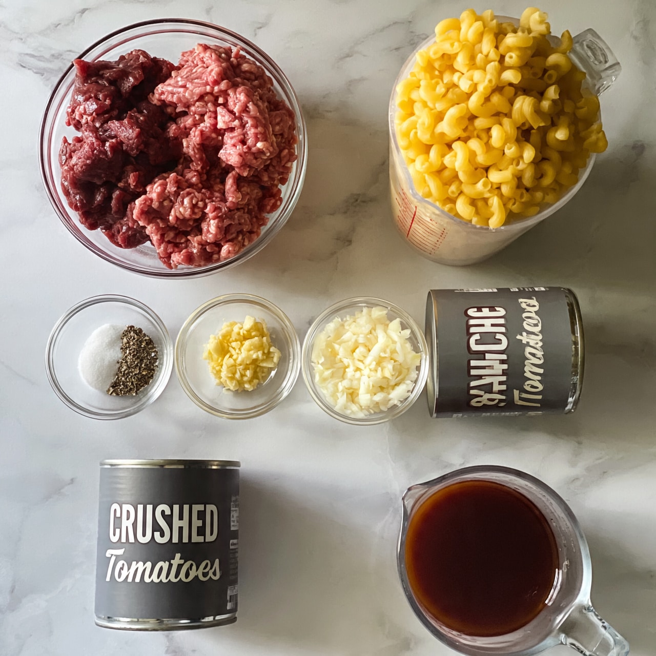 square image The image shows raw ingredients neatly placed on a white marbled surface. There is a clear glass bowl with two types of raw ground meat, one dark red and the other pink, at the top left. Next to it, on the right, is a tall clear measuring cup filled with uncooked yellow elbow macaroni pasta. Below the meat, three small clear bowls hold different ingredients: one bowl has a mix of salt, pepper, and herbs; the middle bowl contains minced garlic; and the last bowl features finely chopped white onions. Two cans of tomato products are visible, one labeled