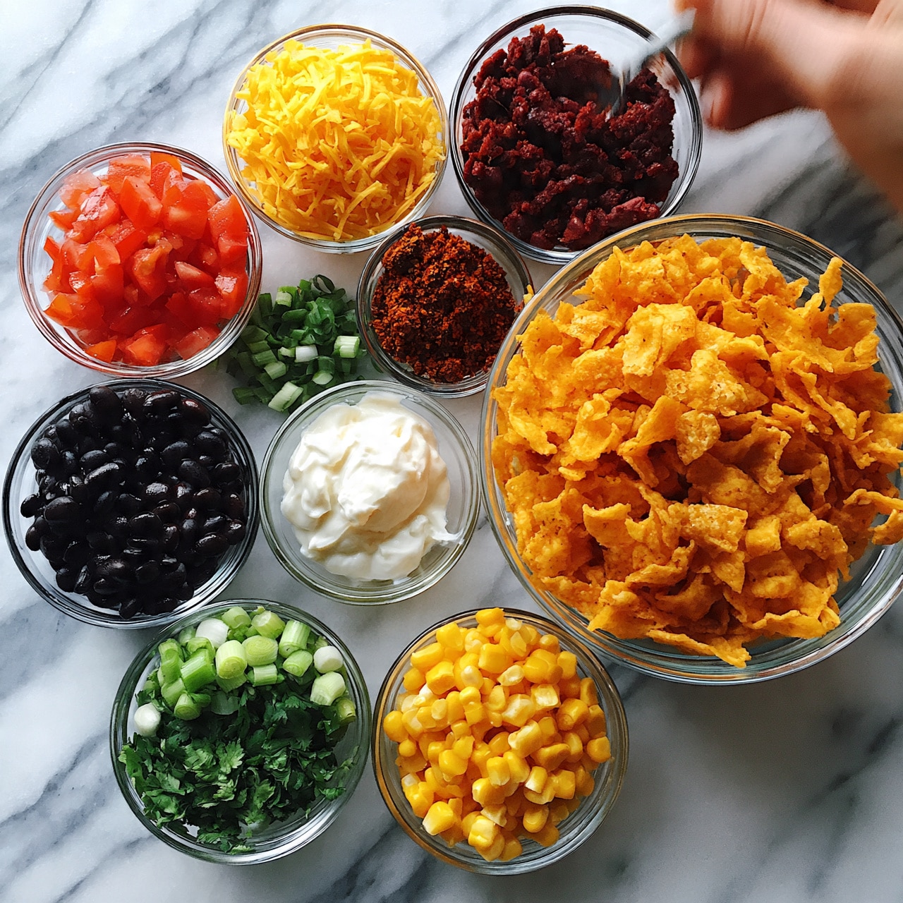square image The image shows an overhead view of several small clear bowls arranged on a white marbled surface, each containing different ingredients for making a dish. The largest bowl holds bright orange crunchy chips in the middle right part of the image. Surrounding it are smaller bowls filled with various colorful items: bright red diced tomatoes, finely shredded yellow cheese, dark black beans, pale white sour cream, chopped green onions, green chopped cilantro, and a deep reddish-brown spice mix. There is also a bowl with finely ground deep red meat or beans and another bowl with bright yellow corn kernels. A woman's hand holding a spoon appears in the top right corner near the brown spice, as if ready to scoop some of the ingredients. The simple and colorful setup offers a clear view of each component in neat, separate bowls. photo taken with an iphone --ar 4:5 --v 7