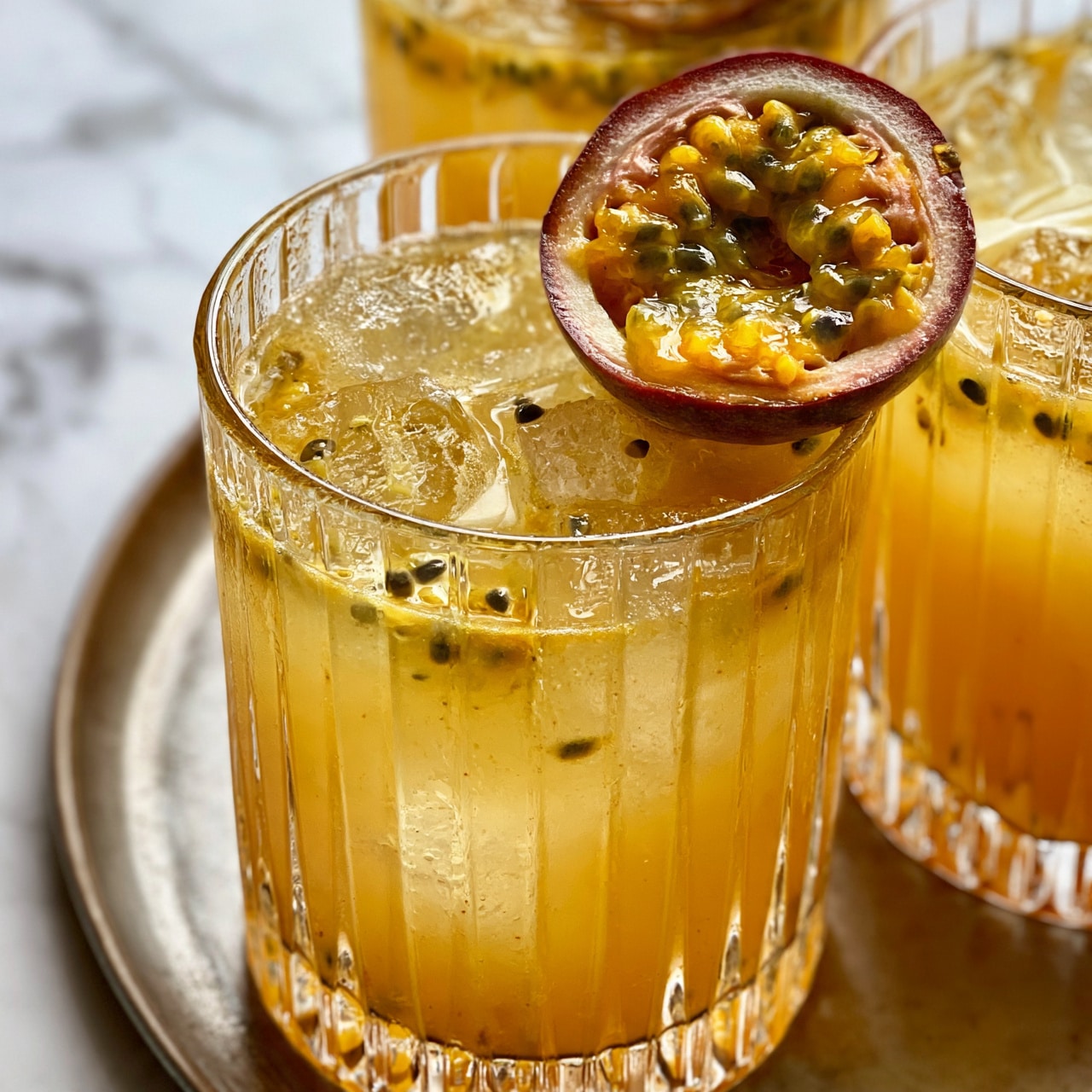 square image The image shows a close-up of a clear glass with vertical ridges filled with a yellowish-orange drink, ice cubes, and small black seeds floating inside. A halved passion fruit with juicy yellow pulp and black seeds rests on the rim of the glass. Around the main glass, there are other glasses filled with similar drinks and ice, placed on a round metal tray. The background is a white marbled surface. The photo taken with an iphone --ar 4:5 --v 7