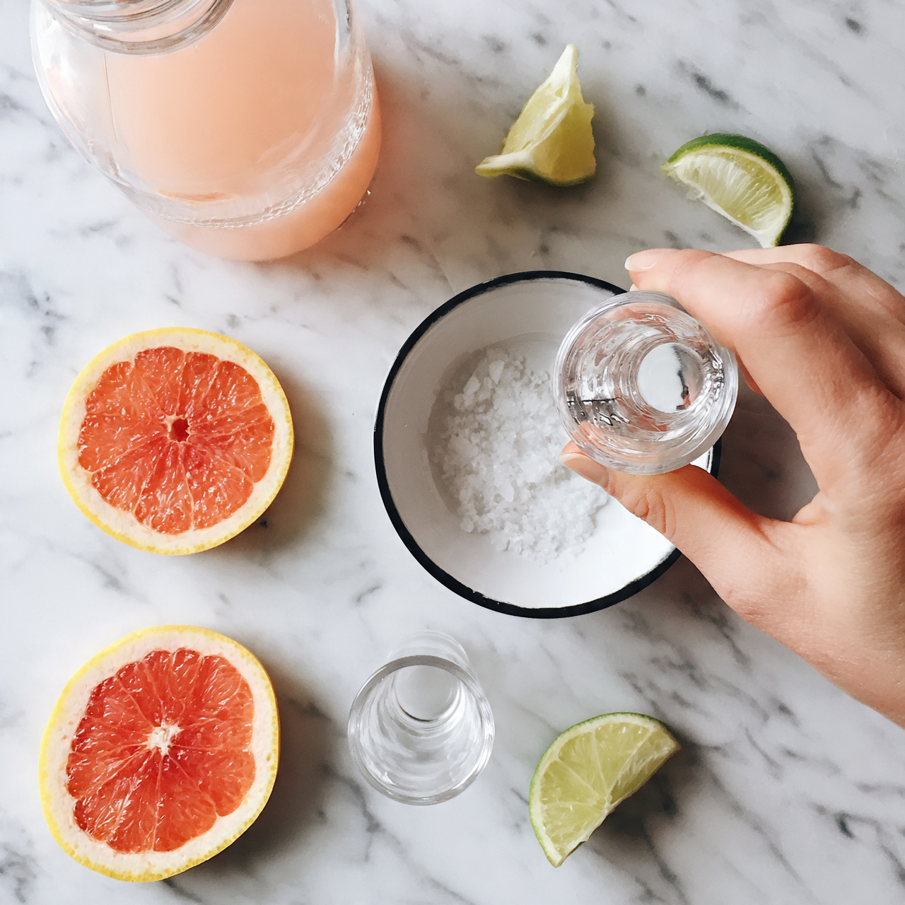 square image On a white marbled surface, there are three slices of pink grapefruit with yellow edges, and two lime wedges with green skin and light green inside, scattered around. Near the top left is a clear glass carafe filled with light pink juice. Near the center is a white round bowl with a black rim containing a small amount of coarse white salt. A woman's hand holds a clear shot glass above the bowl. There is also an empty clear shot glass near the center left of the image. Photo taken with an iphone --ar 4:5 --v 7