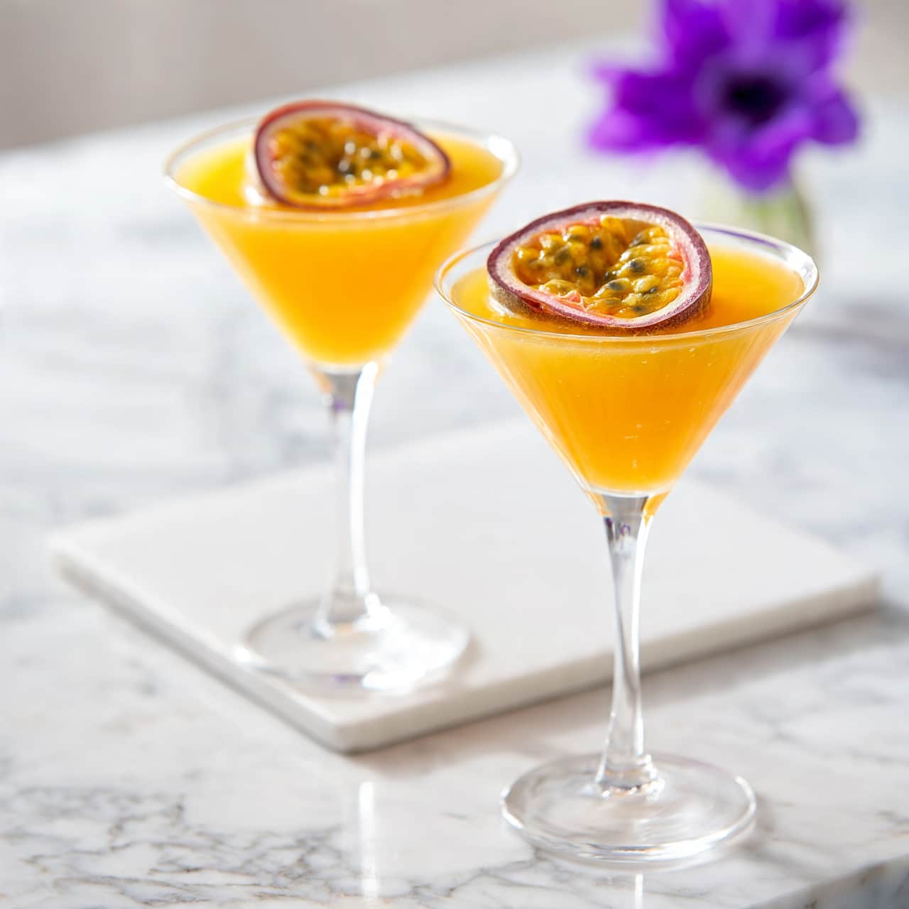 square image Two clear martini glasses filled with bright orange liquid, each topped with a single halved passion fruit showing its yellow pulp and black seeds. The glasses have thin stems and sit on a white marbled surface with a white rectangular cutting board beneath one glass. In the background, there is a blurry purple flower in a small vase, adding a pop of color. The scene is bright and clean, showing a fresh, tropical drink. photo taken with an iphone --ar 4:5 --v 7