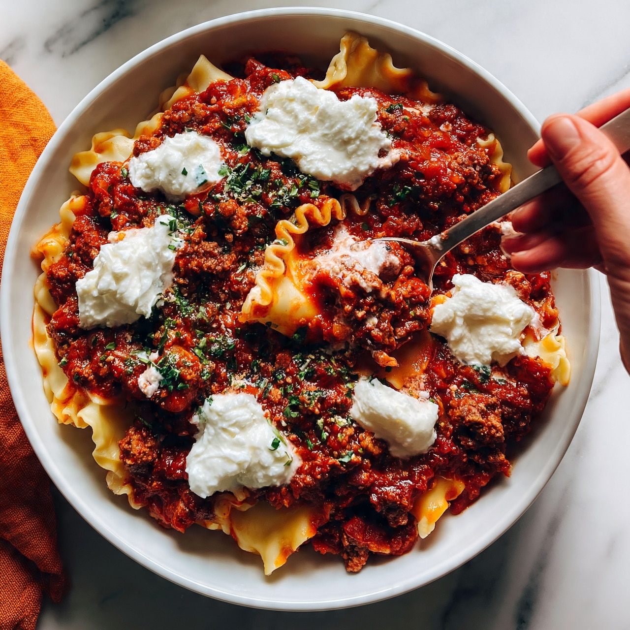 square image A white bowl filled with layered pasta sheets covered in thick, rich red sauce mixed with ground meat, topped with dollops of soft white cheese. The sauce has a textured look with visible bits of meat and herbs, and the cheese appears creamy and smooth. A woman's hand holding a spoon is starting to scoop some from the bowl. The bowl is placed on a white marbled surface with an orange cloth partially visible to the side. Photo taken with an iphone --ar 4:5 --v 7