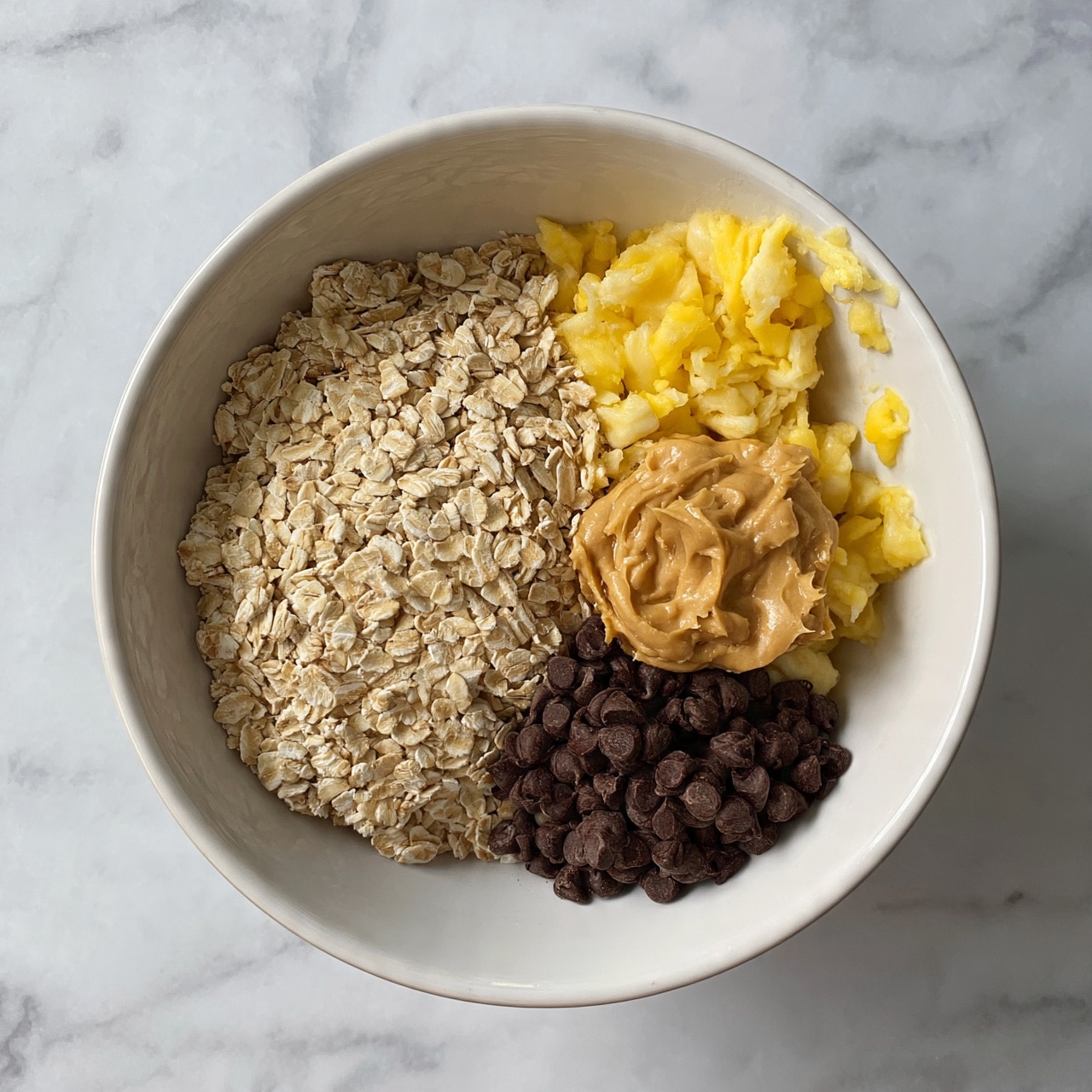 square image A white bowl on a white marbled surface holds four separate sections of ingredients: pale beige dry rolled oats fill the left half, a small heap of dark brown chocolate chips sits at the bottom right, a dollop of creamy light brown peanut butter is placed above the chocolate, and mashed bright yellow banana pieces are scattered on the top right and some edges near the peanut butter. All ingredients show their distinct textures clearly with no mixing photo taken with an iphone --ar 4:5 --v 7