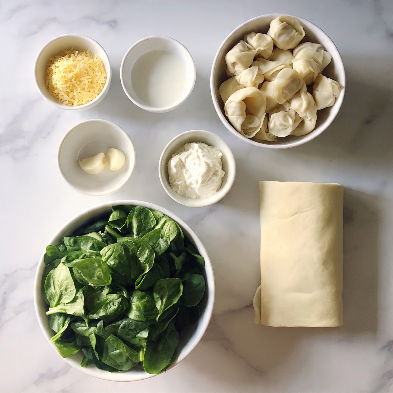 square image The image shows a top view of six bowls and one block item on a white marbled surface. In the top left, a small white bowl holds grated yellow cheese, next to it a small white bowl with a white liquid cream, and to the right, a medium white bowl filled with uncooked wontons. Below these, a small white bowl contains two peeled garlic cloves, beside it a small white bowl with a creamy white soft cheese, and on the bottom left, a large white bowl is full of fresh green spinach leaves. To the right of the large bowl, a block of light beige dough or pastry sheet is placed flat on the marble surface. The photo taken with an iphone --ar 4:5 --v 7