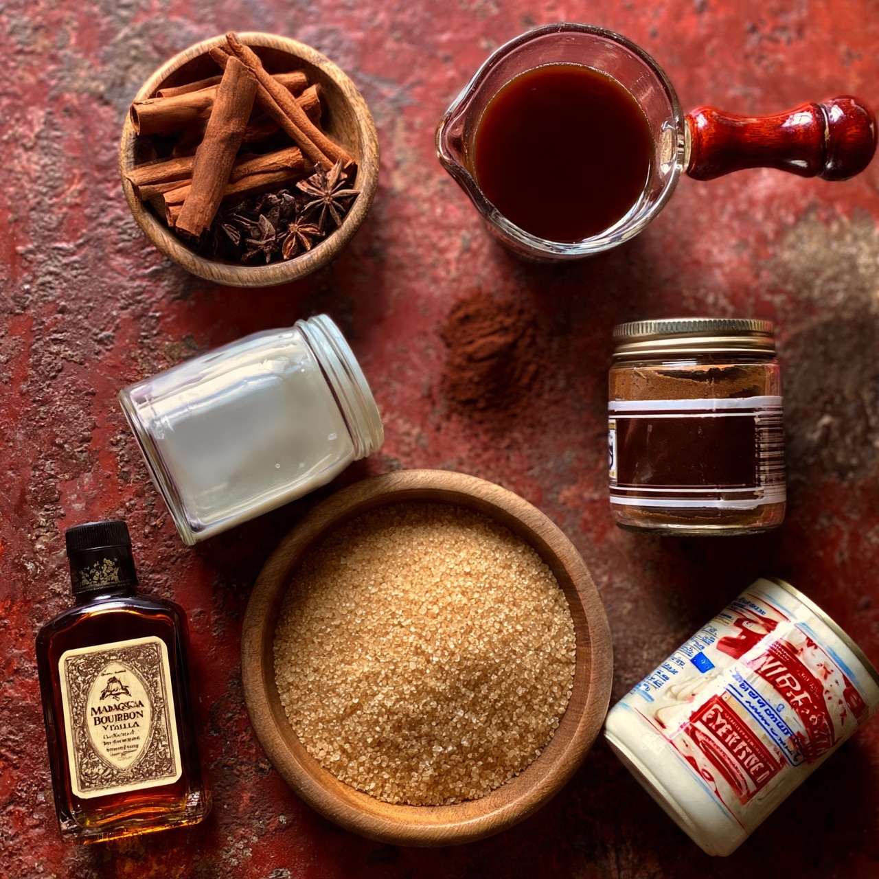 square image A flat lay of ingredients arranged on a rough reddish surface, featuring five main items: at the top left, a small wooden bowl filled with medium brown cinnamon sticks and dark brown star anise pods; to the right, a clear glass measuring cup with a rich dark brown liquid and a polished wooden handle; below that, a glass jar of instant espresso powder with a dark brown and white label; at the bottom left, a tilted clear glass container of white milk; next to it, a round wooden bowl filled with light golden brown raw sugar crystals; bottom left corner shows a dark amber bottle of Madagascar bourbon vanilla extract with a cream label; and at the bottom right, a tilted white can of sweetened condensed milk with red and blue text. All items contrast with the rustic background. photo taken with an iphone --ar 4:5 --v 7