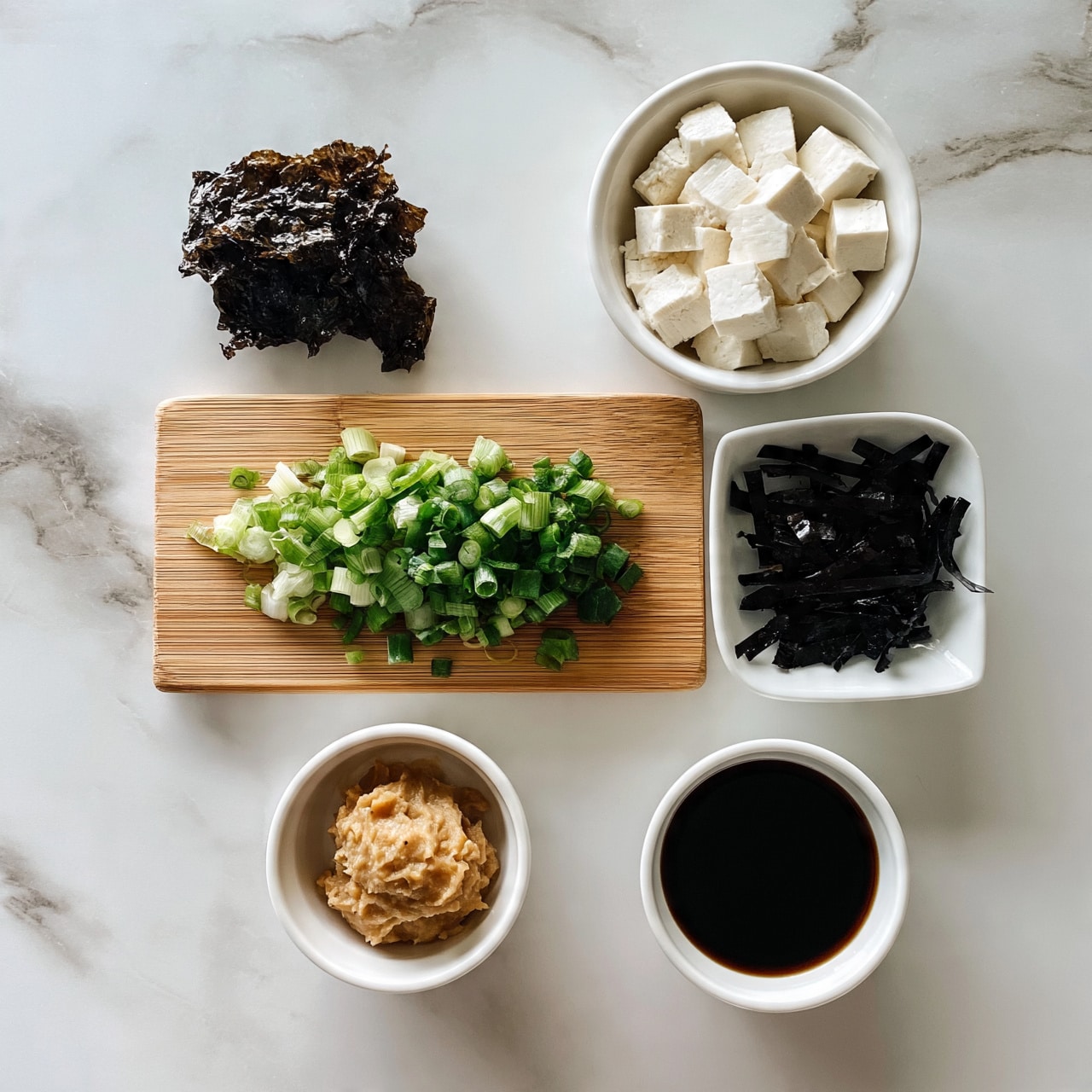 square image The image shows a white marbled surface with six small white containers arranged neatly. In the top left, there is a piece of dark dried seaweed. Next to it on the right, a white bowl filled with small white tofu cubes. Below these, a wooden cutting board holds chopped green onions. To the right of the board, a white dish contains black dried seaweed strips. Below, a small white bowl has light brown miso paste, and next to it, another small white bowl contains dark soy sauce. The arrangement is simple and clean, all items placed clearly on the white marbled surface. photo taken with an iphone --ar 4:5 --v 7
