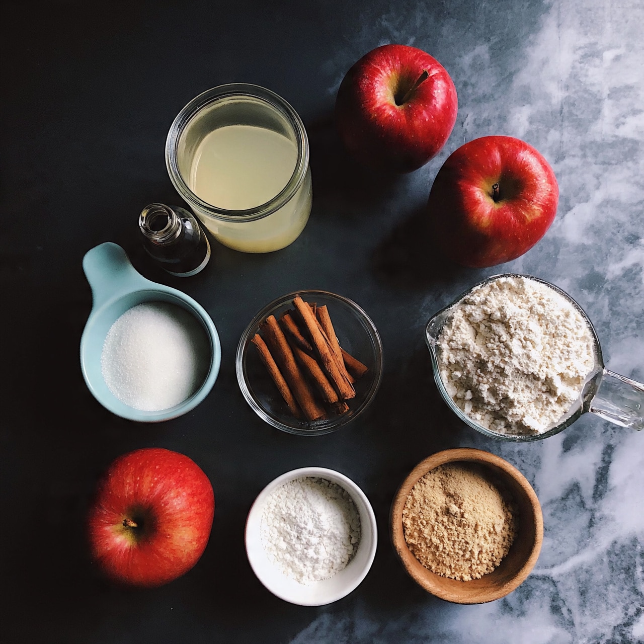 square image The image shows a dark surface with several baking ingredients arranged neatly. There are three red apples placed in different spots. A small glass bowl with brown cinnamon sticks is near the center. A light blue cup filled with white sugar and a measuring cup with white flour are also visible. There is a glass container of pale yellow liquid and a small bottle of vanilla extract. A small white bowl filled with white powder is present, and a wooden bowl filled with a light brown powder is shown. The background is changed to a white marbled texture. photo taken with an iphone --ar 4:5 --v 7
