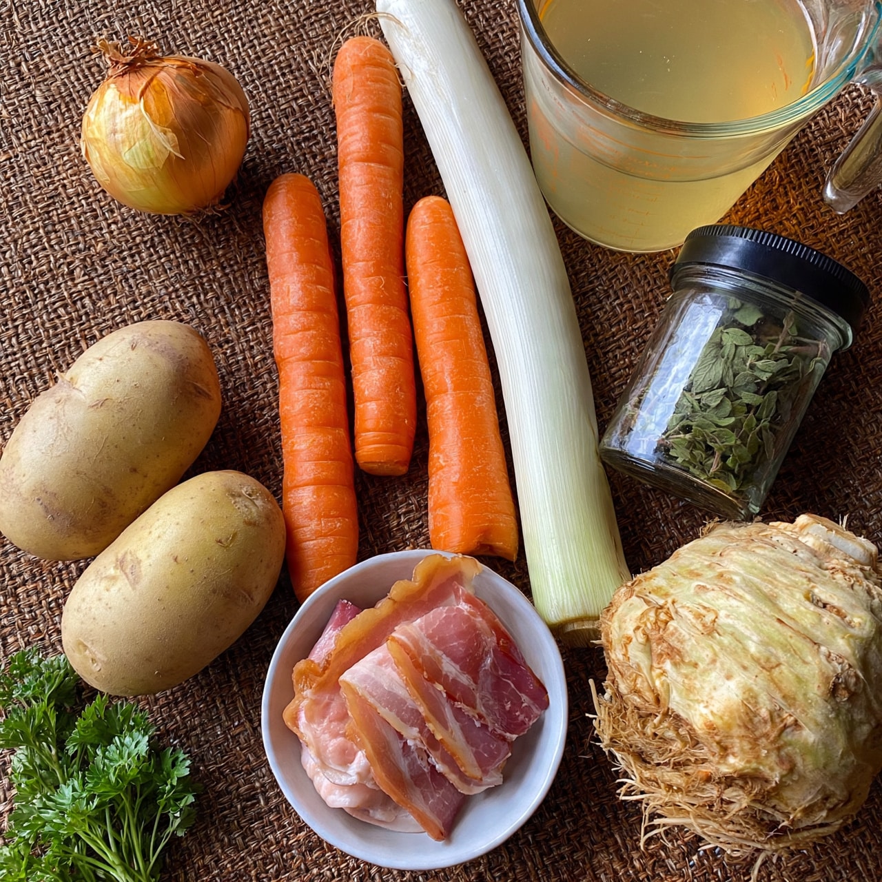 square image The image shows a flat lay of fresh ingredients on a brown woven mat with a few sprigs of parsley at the bottom left. There are two large potatoes with light brown skin near the bottom left and an onion with golden brown skin placed near the top center. Above the onion is a clear measuring cup filled with light yellow broth, positioned to the top right. Three bright orange carrots are arranged vertically near the center, alongside a white leek stalk. A clear jar of marjoram leaves with a black lid is to the right of the carrots. To the bottom right, there is a bulb of celeriac with its rough pale brown skin and root proteins visible. Lastly, in a small white bowl near the bottom center are three slices of smoked bacon with pink meat and fat. Photo taken with an iphone --ar 4:5 --v 7