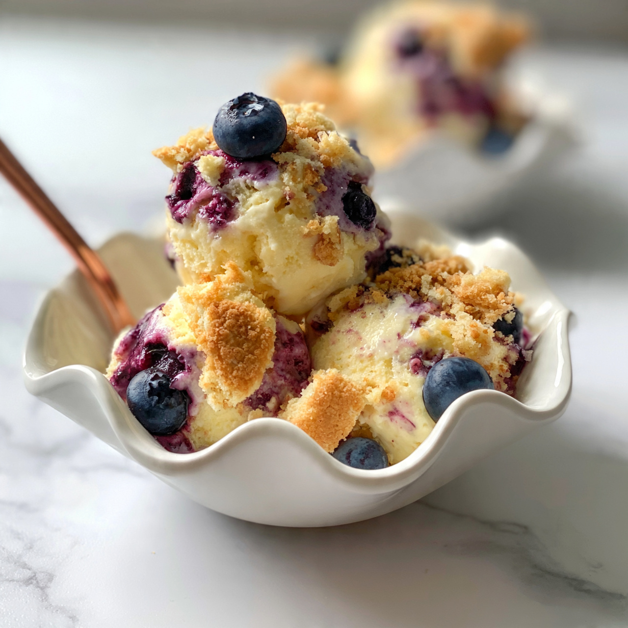 square image The dish is served in a small white bowl with a wavy edge, filled with three scoops of creamy pale yellow ice cream mixed with purple blueberry sauce and dotted with whole blueberries. There are crumbly golden brown biscuit pieces mixed throughout the ice cream, adding texture and color contrast. A copper spoon is placed inside the bowl on the left side, and the bowl sits on a white marbled surface. In the background, another similar bowl with the same dessert is slightly out of focus. Photo taken with an iphone --ar 4:5 --v 7