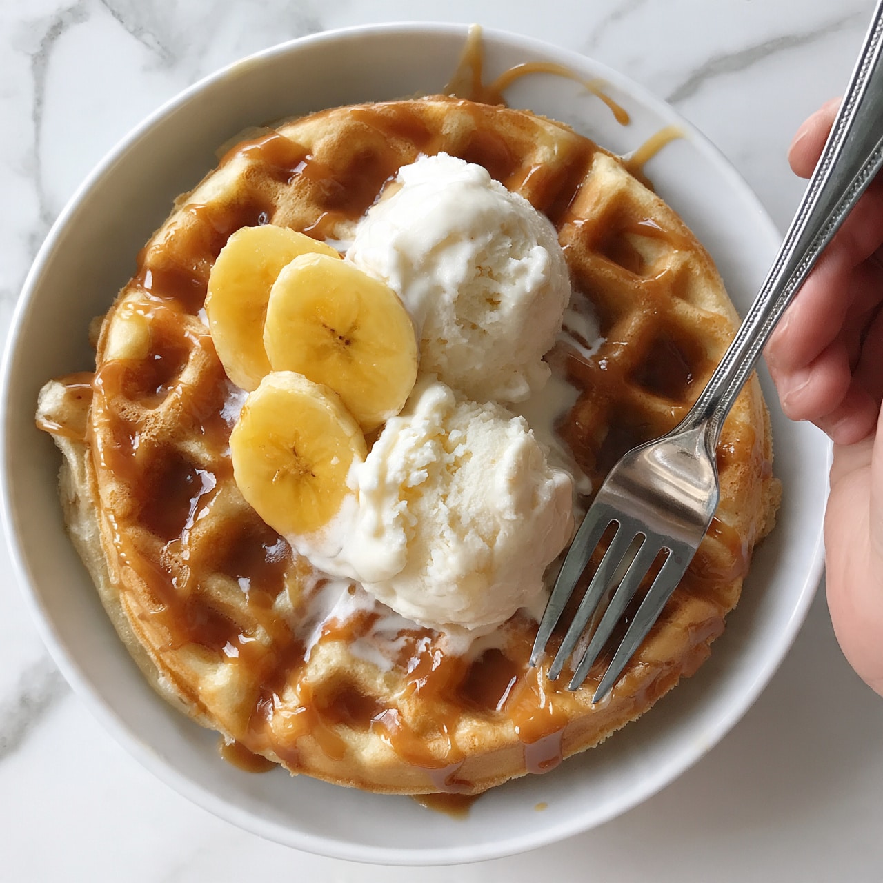 square image A thick golden waffle sits in a white bowl, with a layer of light brown sauce drizzled over it. On top, there are three slices of yellow banana and two scoops of white ice cream, slightly soft and creamy. A silver fork is held by a woman's hand on the right side, with the tines partly inside the waffle. The bowl is placed on a white marbled surface. photo taken with an iphone --ar 4:5 --v 7