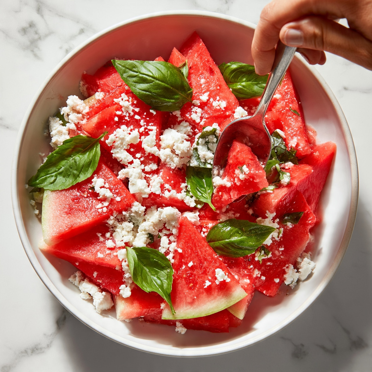 square image A white shallow bowl filled with bright red triangular slices of watermelon arranged in layers, topped with crumbled white cheese scattered unevenly, and fresh green basil leaves placed on top and around the watermelon slices. A metal spoon with a woman's hand holding its handle is scooping some of the salad from the bowl. The bowl sits on a white marbled surface. Photo taken with an iphone --ar 4:5 --v 7