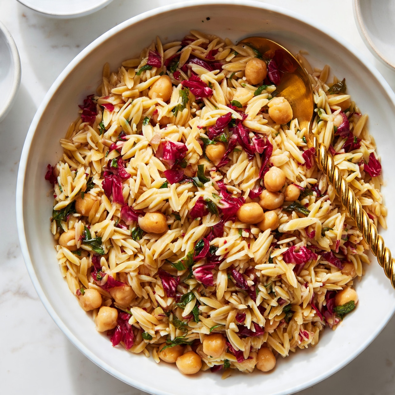 square image The image shows a large white bowl filled with a colorful chickpea and orzo pasta salad. The dish has three main layers: the base layer is light golden orzo pasta mixed evenly throughout. Scattered among the orzo are round, tan chickpeas that add texture. Mixed throughout are small, bright red pieces of radicchio and some green leafy herbs, adding pops of deep red and green colors. A golden twisted spoon is placed inside the bowl, resting on the salad. The bowl sits on a white marbled surface with other smaller white bowls partially visible in the background. The lighting is bright and natural, creating a fresh and appetizing look. photo taken with an iphone --ar 4:5 --v 7