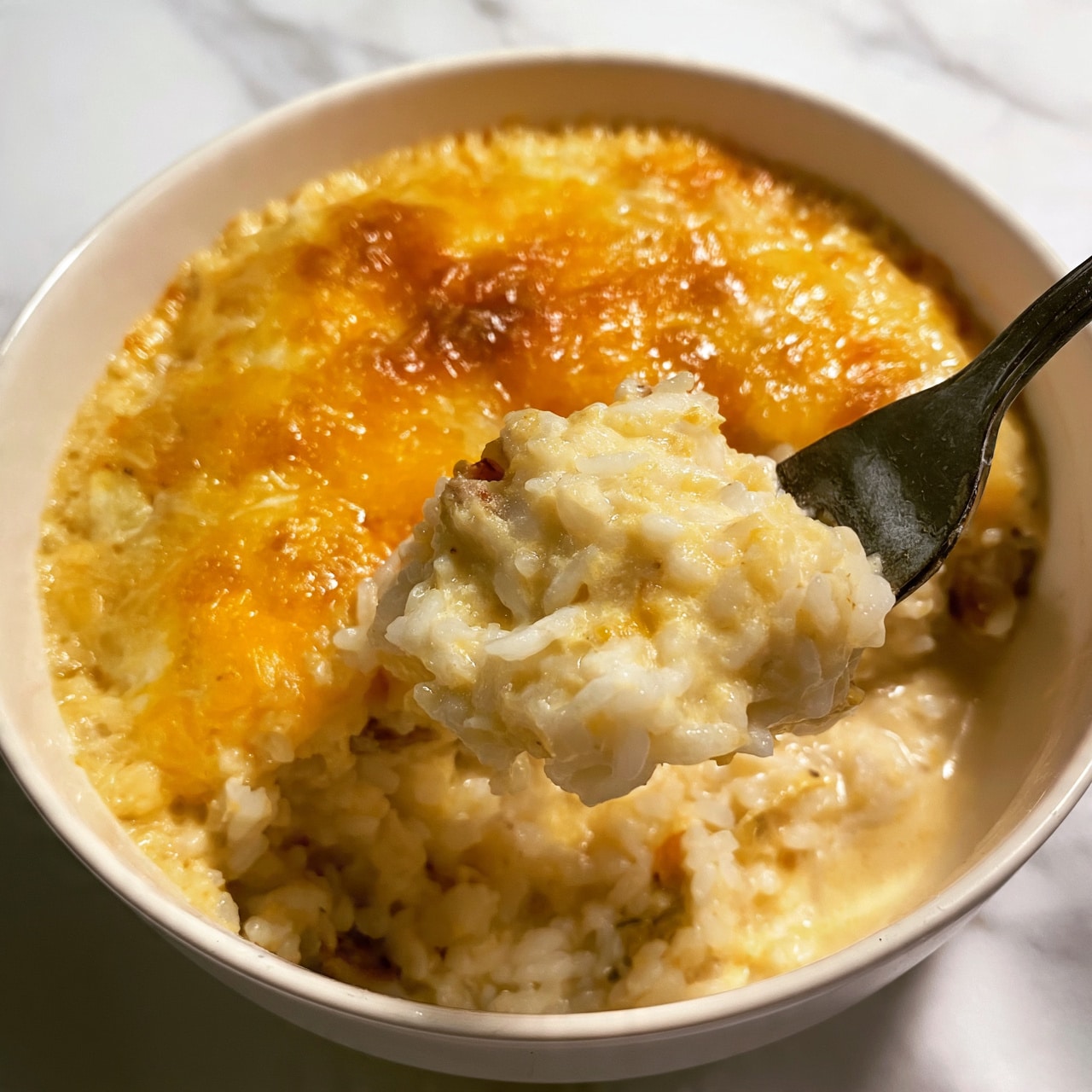 square image A close-up view of a rice dish inside a white bowl with a creamy golden layer of melted cheese or sauce on top, showing a slightly browned and textured surface. The dish has two visible layers: the top layer is the cheesy golden crust that looks soft and shiny, while the bottom layer is a mix of cooked white rice and small bits of vegetables or meat in a creamy sauce. A metal fork is lifting a scoop of the rice mixture, revealing the soft, moist texture inside. The bowl sits on a white marbled surface, and the overall look is warm and comforting. Photo taken with an iphone --ar 4:5 --v 7