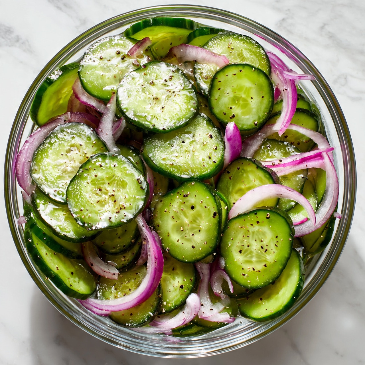 square image A close-up of a glass bowl filled with thin round slices of fresh cucumber and thin strips of purple-red onion mixed together. The cucumber slices are bright green with dark edges and small black pepper flakes on top, showing a shiny, moist texture. The onion strips add a contrast of pale purple and white layers curling around and underneath the cucumber slices. The bowl sits on a white marbled surface. photo taken with an iphone --ar 4:5 --v 7