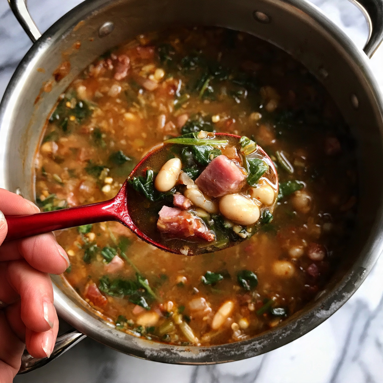 square image A large silver metal pot filled with thick soup containing pieces of ham, beans, spinach, and other small vegetables in a brown broth. A red spoon is held by a woman's hand, scooping some of the colorful soup showing pink ham, white beans, and green spinach clearly. The background is a white marbled surface. photo taken with an iphone --ar 4:5 --v 7