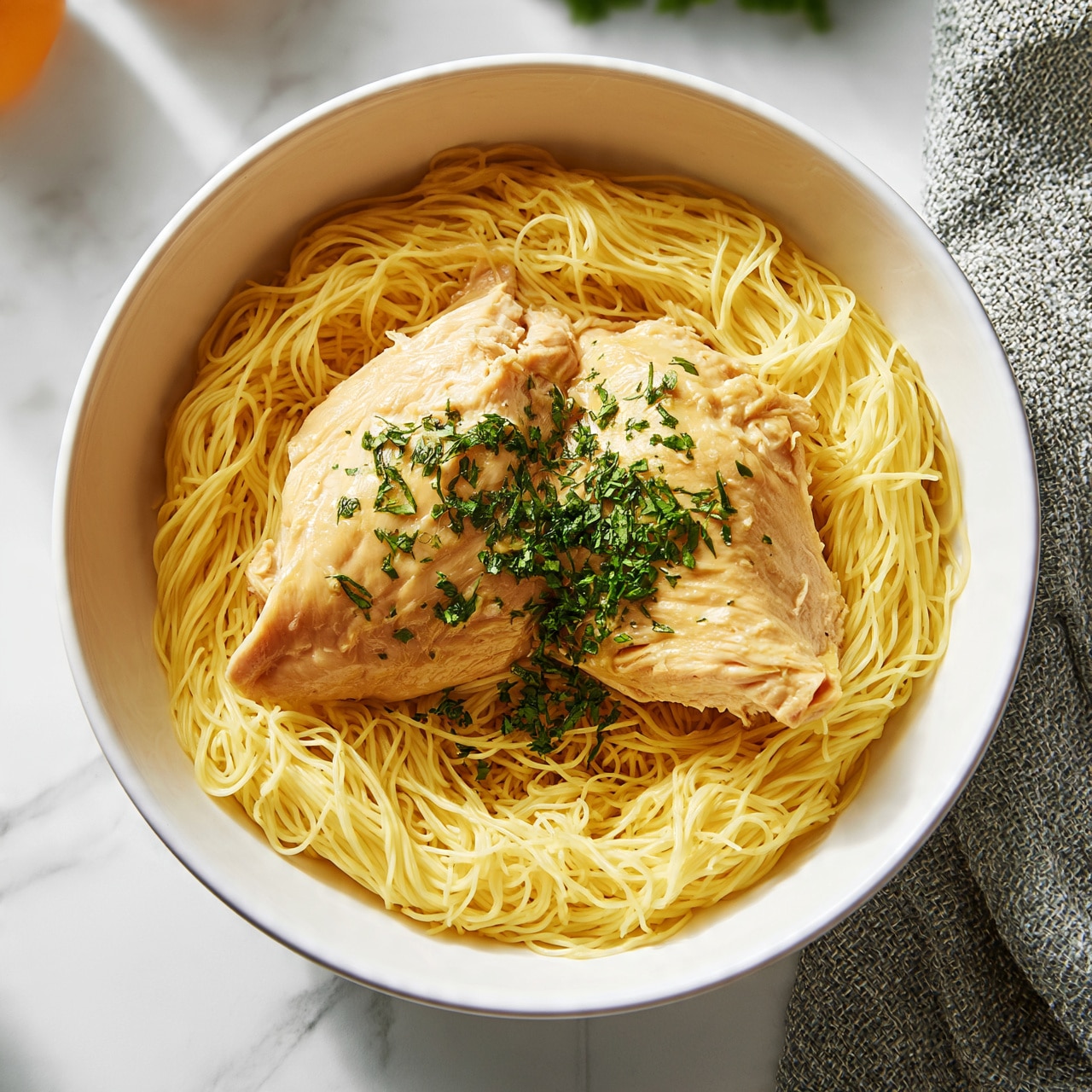 square image A white bowl holds a nest of thin, yellow noodles arranged in a circular pattern, forming the base layer. On top of the noodles, there are two pieces of cooked chicken placed side by side, with a light golden color and smooth texture. The chicken is sprinkled with chopped green herbs, adding a fresh, vibrant contrast. The bowl rests on a white marbled surface with a textured gray cloth nearby and some orange and green objects partially visible in the background. Photo taken with an iphone --ar 4:5 --v 7