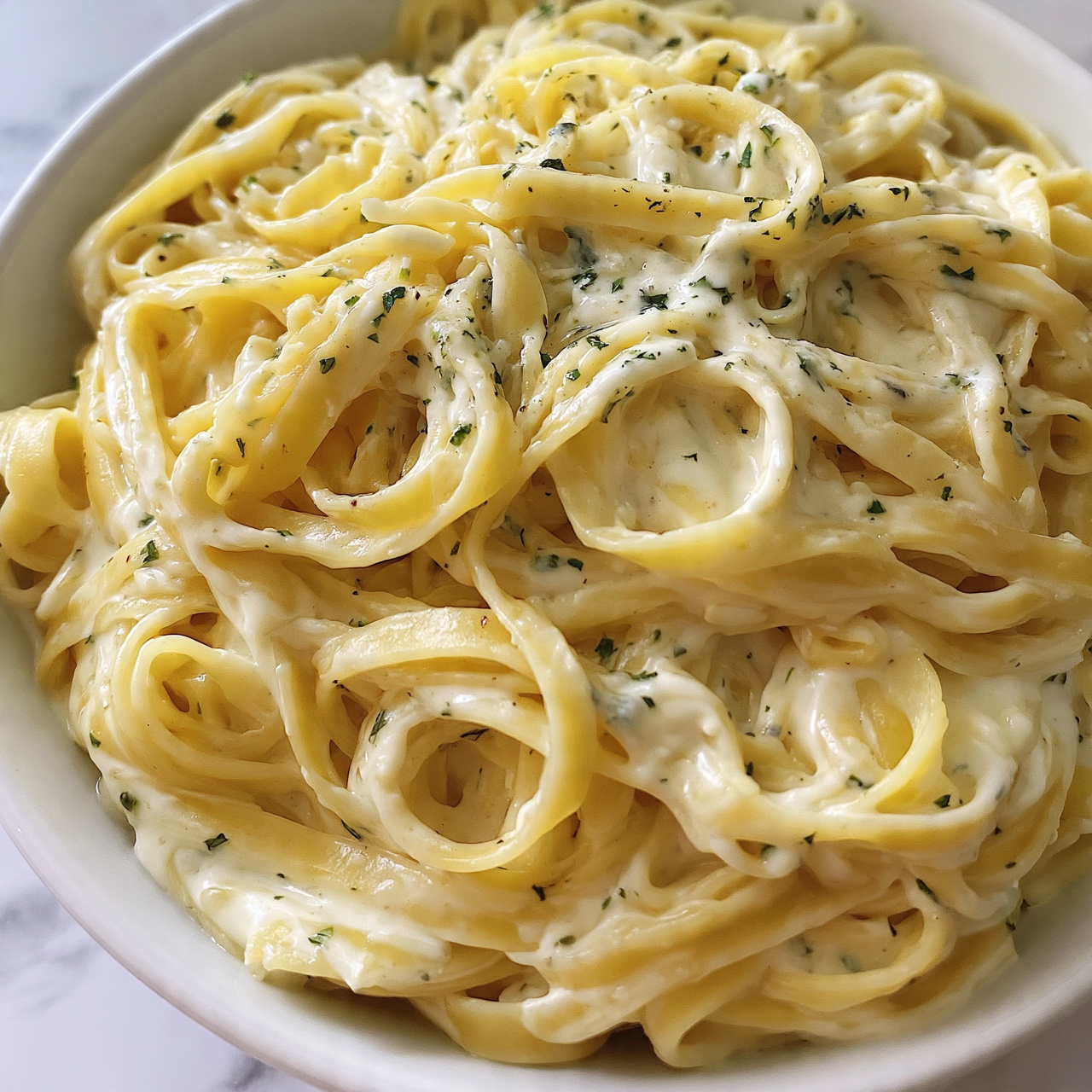 square image A close-up view of a white bowl filled with creamy fettuccine pasta, showing multiple thick, flat noodles tangled together. The noodles are light yellow, mixed with a smooth sauce that looks creamy white with small green herb specks sprinkled all over. The pasta fills the bowl almost to the top, and the white marbled surface is faintly visible around the edges. photo taken with an iphone --ar 4:5 --v 7