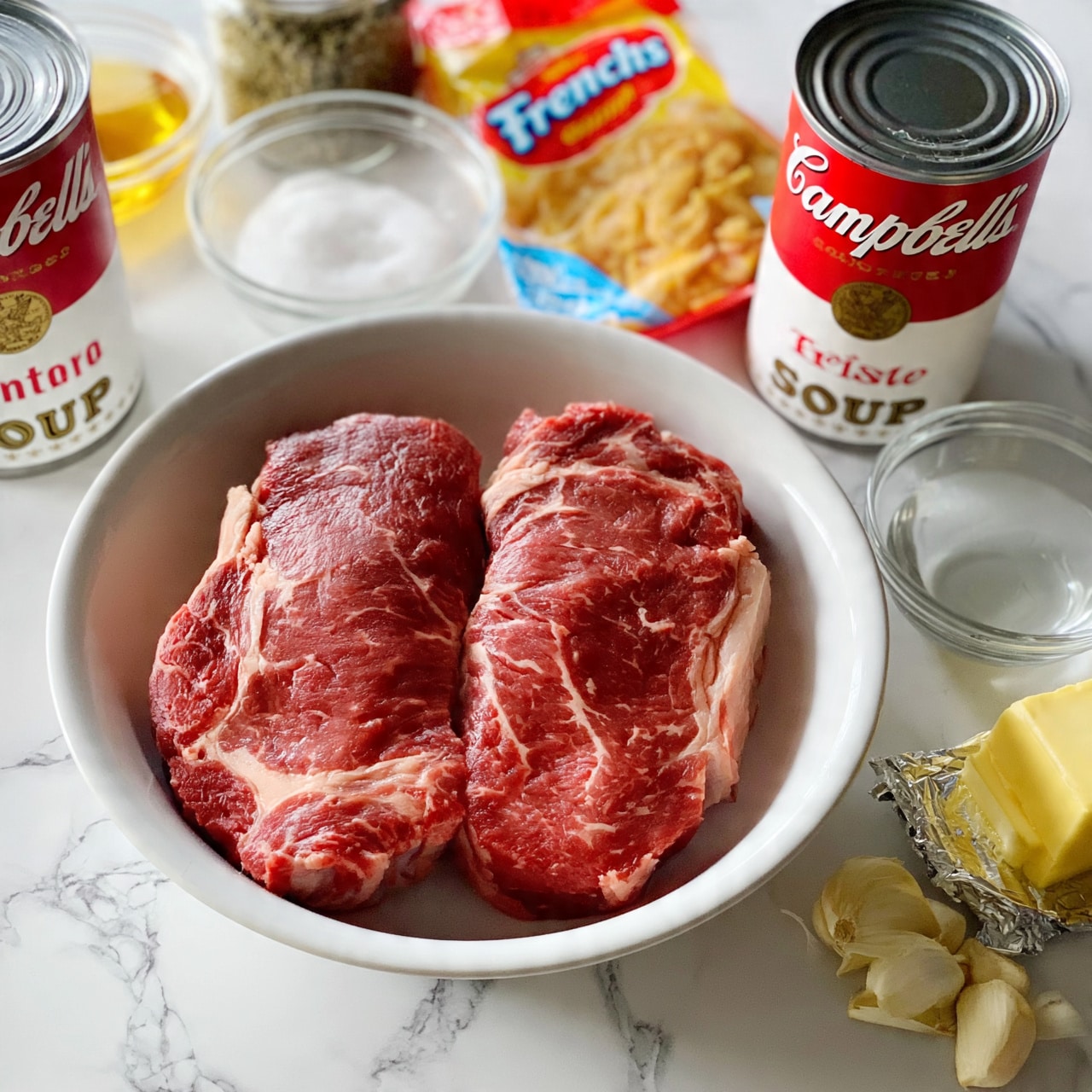 square image A white bowl is placed on a white marbled surface, holding two thick red raw beef steaks with visible white marbling. Around the bowl are two red and white Campbell's soup cans, a clear glass bowl filled with white salt, a small glass bowl with a golden-yellow liquid, a jar of dried herbs, a package of French's crispy fried onions, a small glass bowl with clear water, a stick of yellow butter with wrapper partially open, and several fresh garlic cloves. All items are neatly arranged and clearly visible, creating an organized cooking setup. Photo taken with an iphone --ar 4:5 --v 7