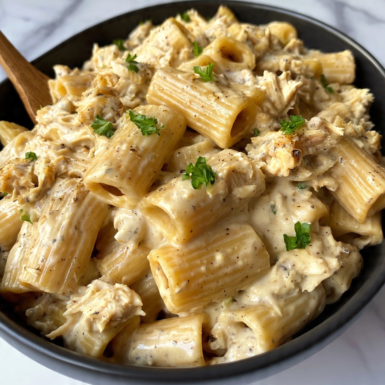 square image The image shows a close-up of a black oval bowl filled with rigatoni pasta coated in a creamy, beige sauce mixed with shredded chicken. The sauce has a smooth, slightly speckled texture from herbs and spices. Small green parsley leaves are scattered on top for garnish. A wooden spoon is partially submerged in the pasta on the left side. The bowl sits on a white marbled surface photo taken with an iphone --ar 4:5 --v 7
