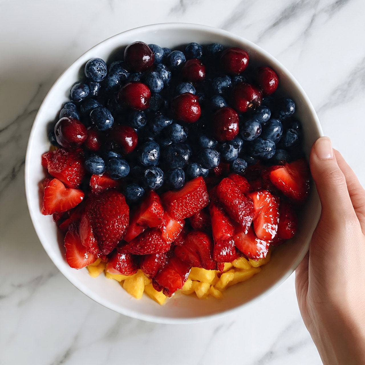 square image A white bowl filled with a colorful fruit salad sits on a white marbled surface. The salad has three main layers: the bottom layer shows small pieces of peeled yellow mango visible through the fruit on top; the second layer is made of sliced red strawberries covering much of the bowl; scattered on top are deep blue blueberries and small whole dark red cherries. The fruits are fresh and shiny, with a mix of smooth and slightly bumpy textures. A woman's hand is holding the edge of the bowl gently from the right side. photo taken with an iphone --ar 4:5 --v 7