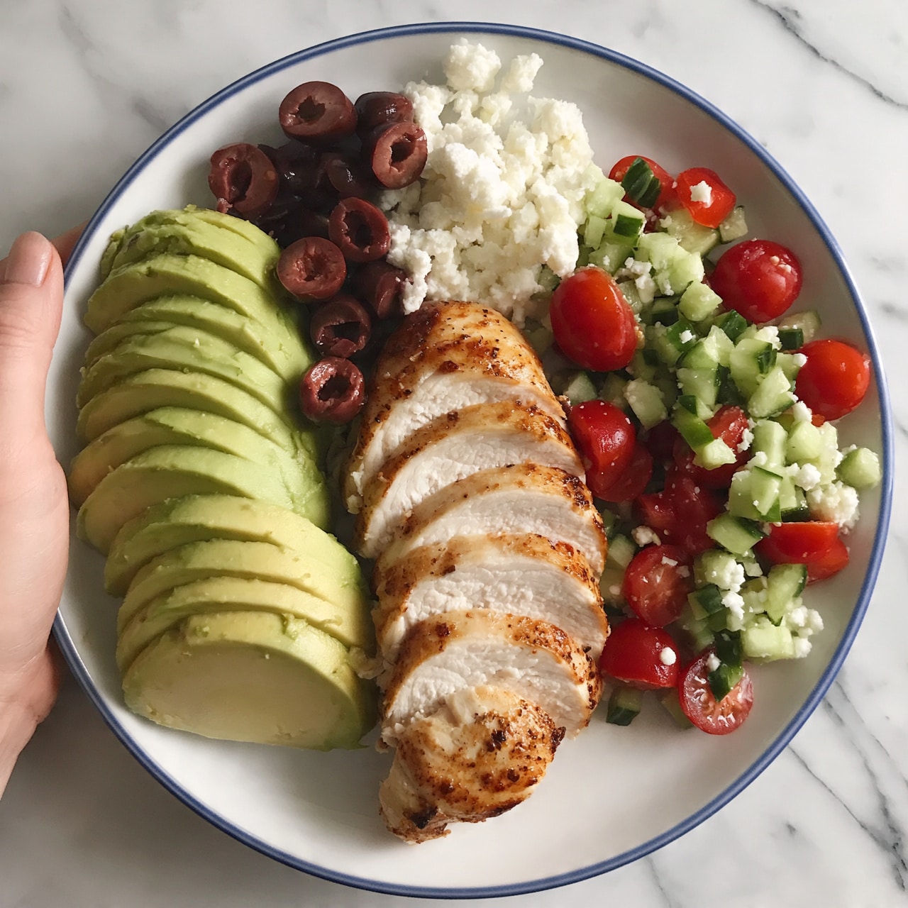 square image The dish is arranged on a white plate with a blue rim, placed on a white marbled surface. In the center, there is a sliced piece of cooked chicken, light golden brown, with thin even slices showing the tender inner texture. Around the chicken, from left to right, are neat sections of creamy avocado slices in light green, small dark reddish-brown olives with pitted centers, white crumbly cheese scattered on top, and a colorful salad made of small diced green cucumbers and bright red cherry tomatoes, mixed with bits of white cheese. A woman's hand is holding the edge of the plate, visible on the left side. The image is bright and clear, showing the fresh textures and colors of the ingredients. photo taken with an iphone --ar 4:5 --v 7
