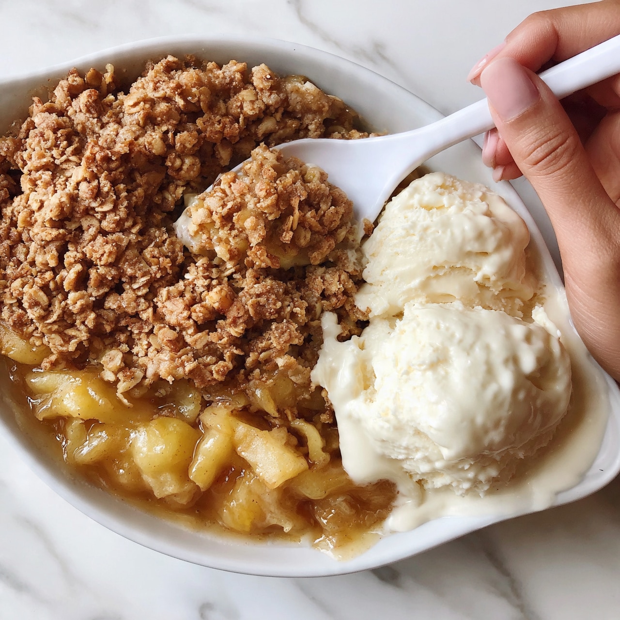 square image The image shows a close-up of a baked apple dessert served in a white dish. The bottom layer is soft apple pieces cooked to a golden color, surrounded by a thick, sticky syrup. On top is a crumbly, brown oat mixture that adds texture and crunch. To the side, there are two scoops of creamy vanilla ice cream with a smooth, slightly melting surface. A woman's hand is holding a white spoon scooping into the warm apple crumble and ice cream. The background is a white marbled surface. photo taken with an iphone --ar 4:5 --v 7