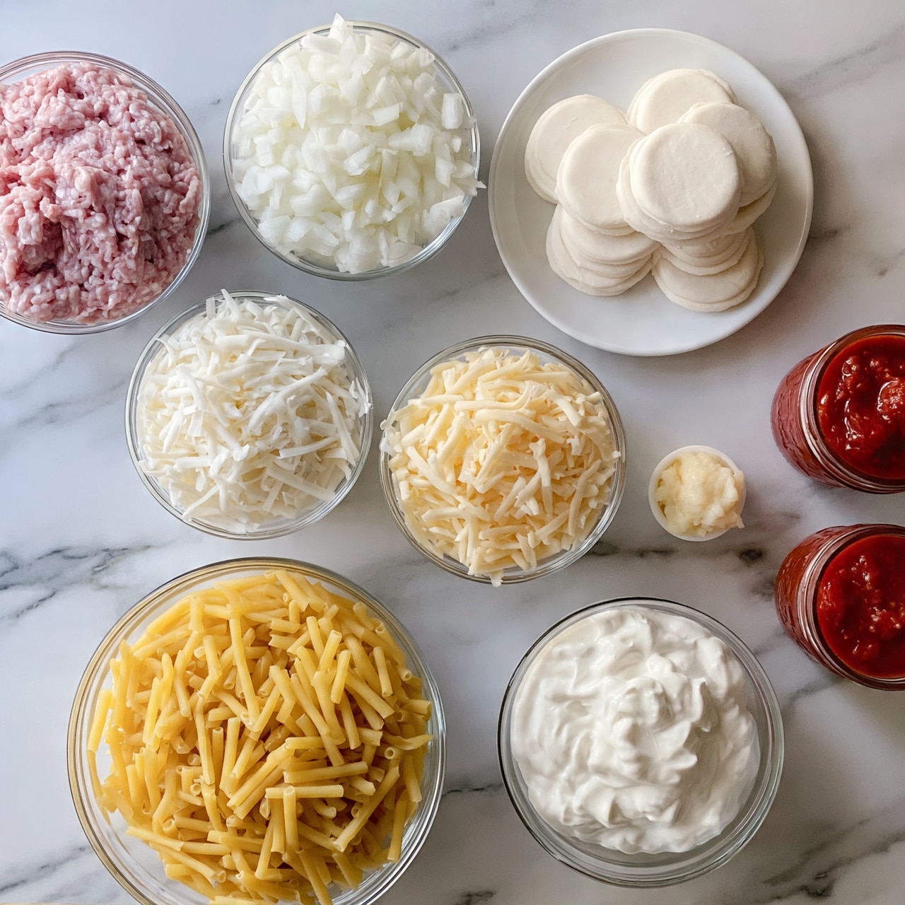 square image The image shows a white marbled surface with several clear bowls and small plates arranged neatly. Starting from the top left, there's a bowl filled with raw ground meat that is light pink. To the right of it is a bowl of finely chopped white onions. Next to the onions are small, round, white dough circles stacked on a white plate. Below them, in the middle, there are two bowls of shredded cheese – one is white and the other is light yellow. Beside the cheeses is a very small bowl of minced garlic, pale off-white in color. On the bottom left, there is a large bowl of uncooked, yellow pasta. To the right of the pasta, a bowl of white sour cream is placed. Lastly, on the right side, two small glass jars filled with red tomato sauce sit on the surface. photo taken with an iphone --ar 4:5 --v 7