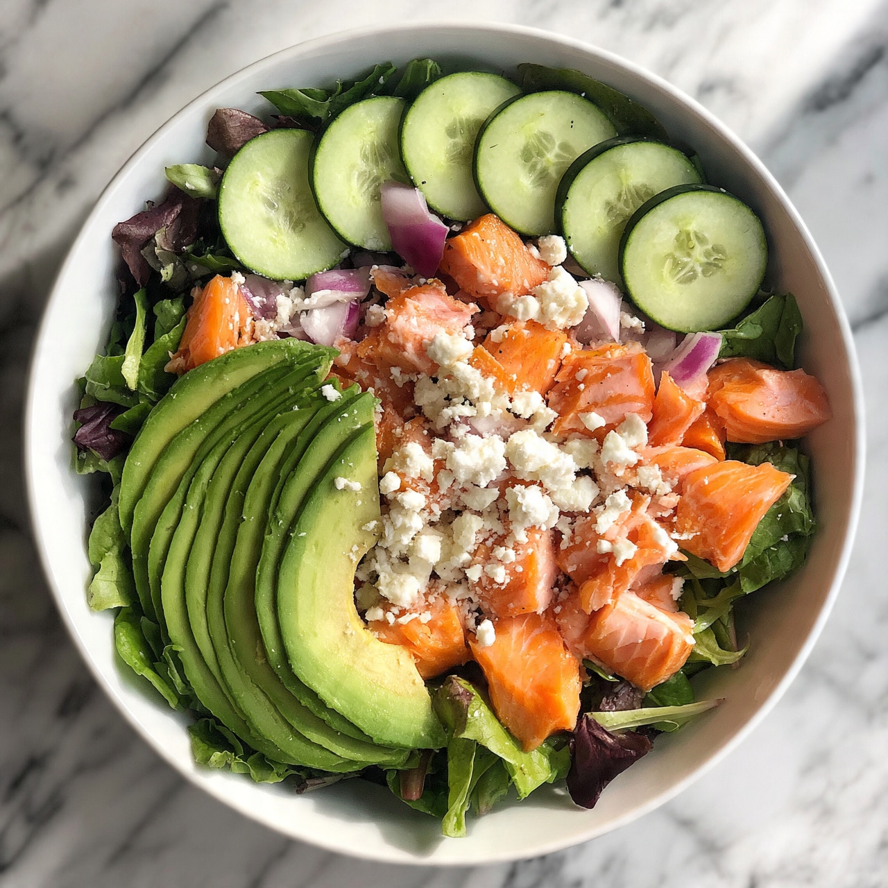 square image A white bowl filled with a fresh salad on a white marbled surface. The salad has several layers: a base layer of mixed green leaves, thin slices of avocado arranged in a curved pattern on one side, chunks of cooked salmon spread throughout, thin half-moon slices of cucumber, small pieces of red onion, and sprinkled white feta cheese on top. The colors are green, pink-orange, white, and purple, creating a bright and healthy look. Photo taken with an iphone --ar 4:5 --v 7