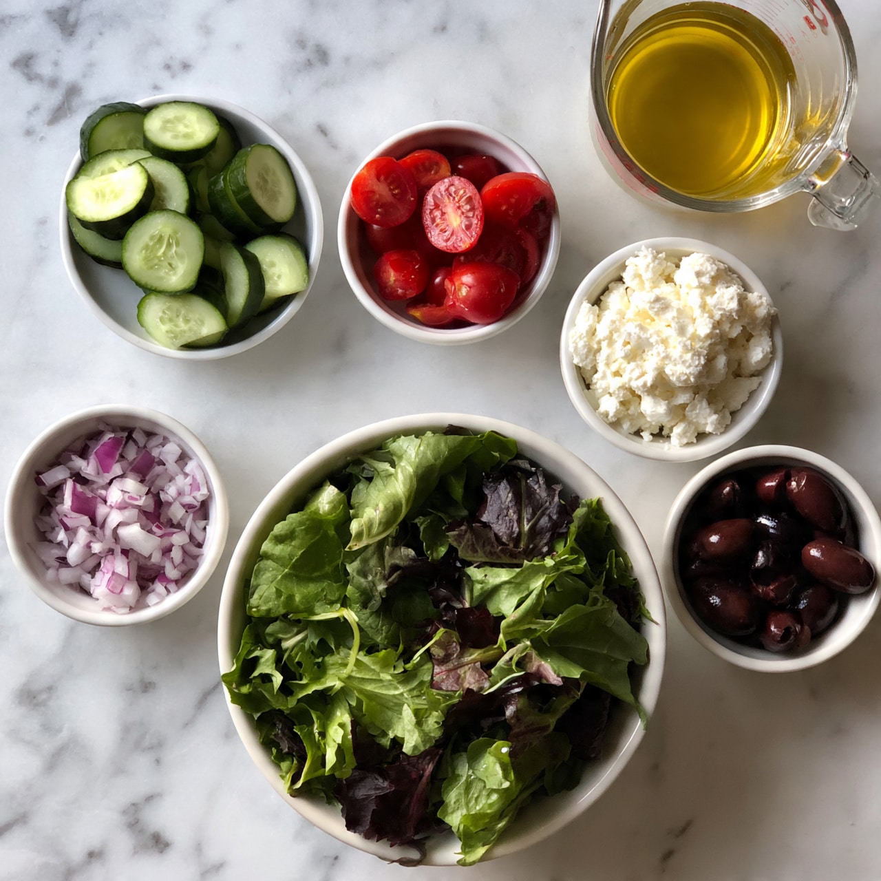 square image The image shows six small white bowls and a clear glass measuring cup arranged on a white marbled surface. The largest bowl at the center bottom is filled with mixed fresh green salad leaves. Above and around it are five small white bowls containing sliced cucumbers, chopped red onions, halved cherry tomatoes, white crumbled cheese, and dark olives. The clear glass measuring cup holds a yellow liquid, likely a dressing, and is placed at the top right. The bowls create a neat, organized layout with colorful, fresh ingredients. Photo taken with an iphone --ar 4:5 --v 7square image