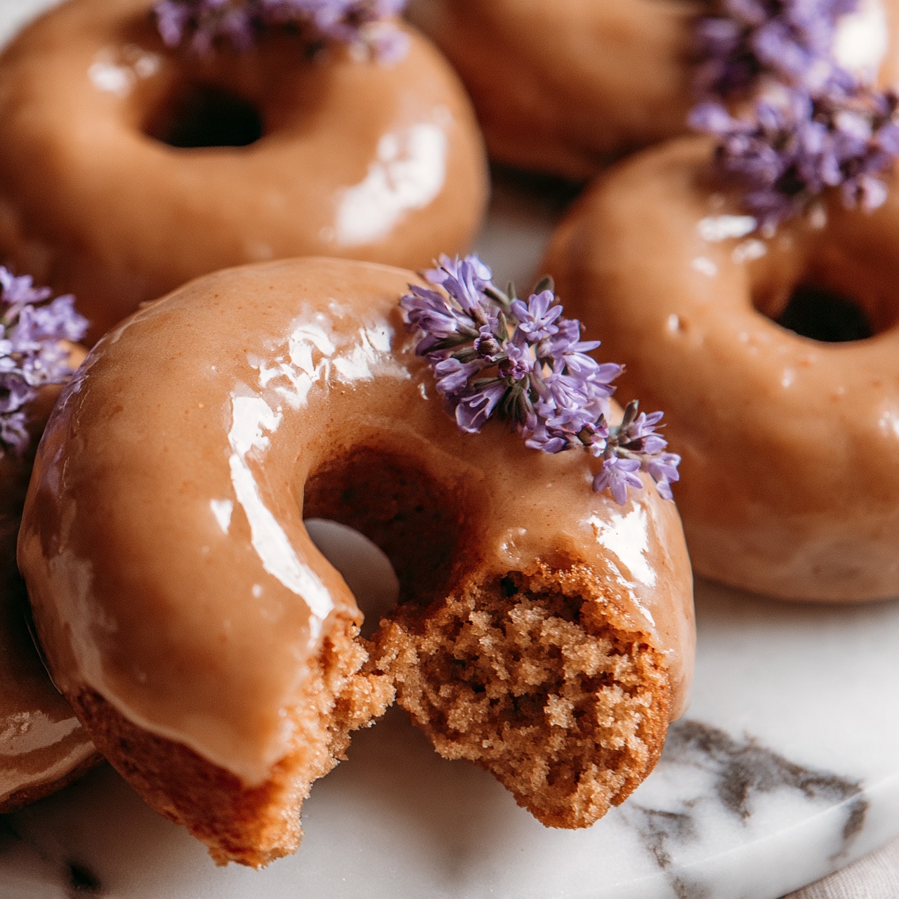 square image The image shows a close-up of several doughnuts with smooth light brown glaze covering each one. They are arranged closely on a white marbled surface, and one doughnut has a bite taken out, showing a soft, fluffy inside that is darker brown. Small, delicate purple flowers are placed on top of some doughnuts, adding a pop of color. The doughnuts have a shiny, smooth texture on the glaze, reflecting light softly. photo taken with an iphone --ar 4:5 --v 7