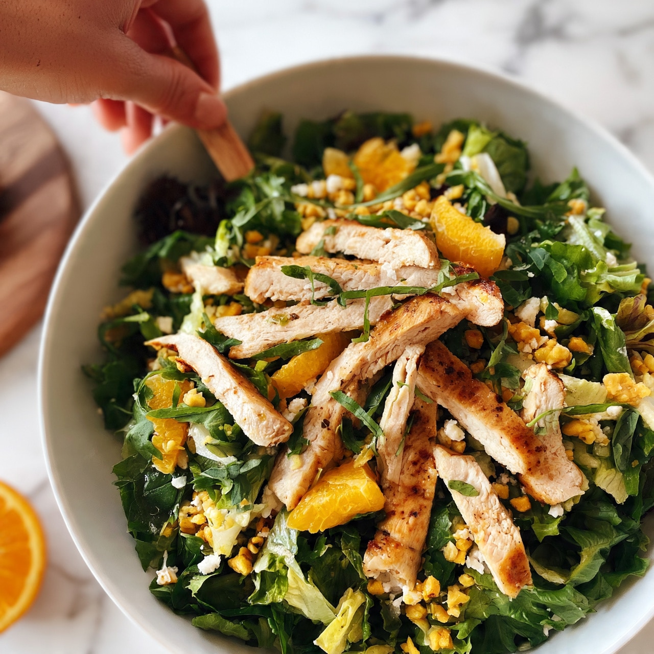 square image A white bowl filled with a fresh salad sits on a white marbled surface. The salad has multiple layers including a base of dark green leafy lettuce mixed with lighter green pieces, topped with thin strips of light brown grilled chicken. Scattered on top are bright orange segments and small white pieces of cheese. Small pieces of a yellow nut or seed add texture, and thin green herb leaves are spread across the salad. A woman's hand holds a small piece of wood near the top left of the bowl. An orange slice and part of a wooden board are visible on the white marbled surface nearby. photo taken with an iphone --ar 4:5 --v 7