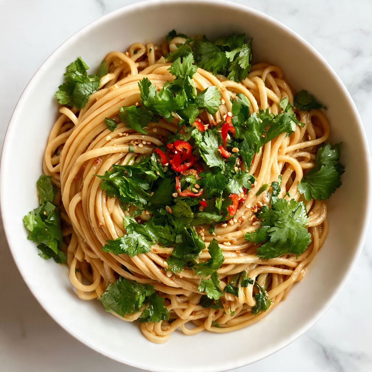 square image A white bowl filled with thick, light brown noodles arranged in a loose swirl, topped with fresh, bright green cilantro leaves scattered across the surface. Thin slices of red chili pepper add small pops of red color on top, while bits of sesame seeds are sprinkled lightly over the noodles. The noodles appear shiny with sauce, showing a smooth texture, and some green herbs are mixed in. The bowl sits on a white marbled surface. photo taken with an iphone --ar 4:5 --v 7
