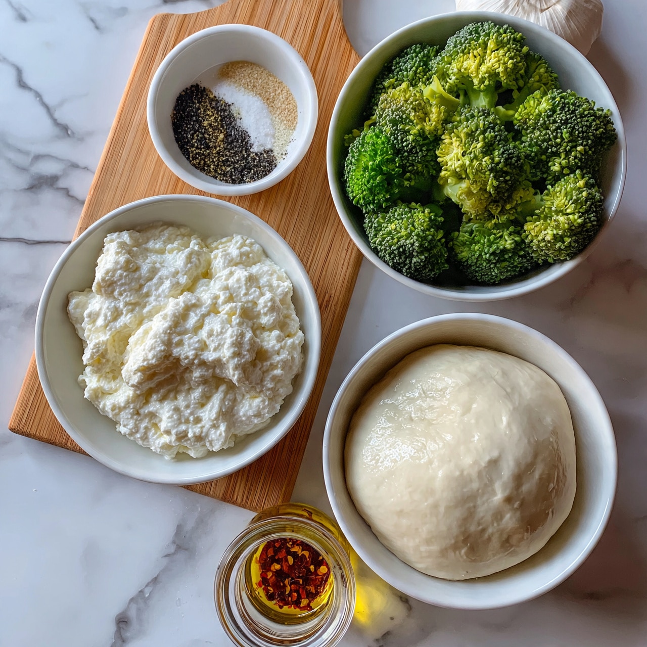 square image The image shows several ingredients arranged neatly on a white marbled surface: a wooden board holds a white bowl filled with thick, creamy white cheese and a small white bowl with mixed spices in layers of black, white, and light brown. Next to the board, a white bowl contains bright green, fresh broccoli florets, and another white bowl holds a smooth, round ball of dough with a slightly shiny texture. Below these, there is a clear glass container with golden oil and red flakes inside. A whole garlic bulb sits near the top right corner. photo taken with an iphone --ar 4:5 --v 7