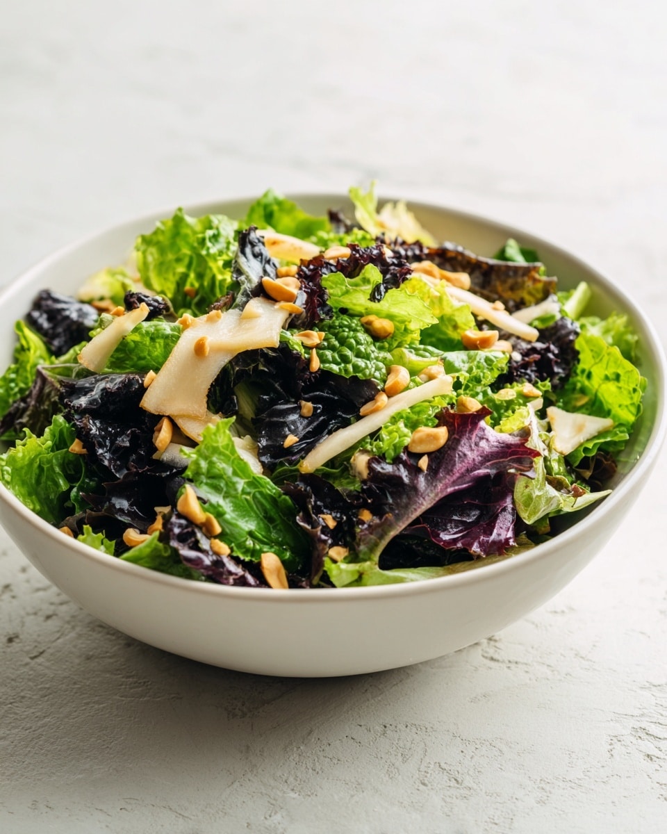 A white bowl filled with a fresh salad on a white marbled surface. The salad has multiple layers including chopped green lettuce, dark purple leafy greens, and thin pale beige strips that look like mushrooms or squid. On top, there are small light tan pieces that look like toasted corn or nuts, scattered evenly. The textures vary from crunchy greens to soft-looking strips and crunchy small pieces. Photo taken with an iphone --ar 4:5 --v 7