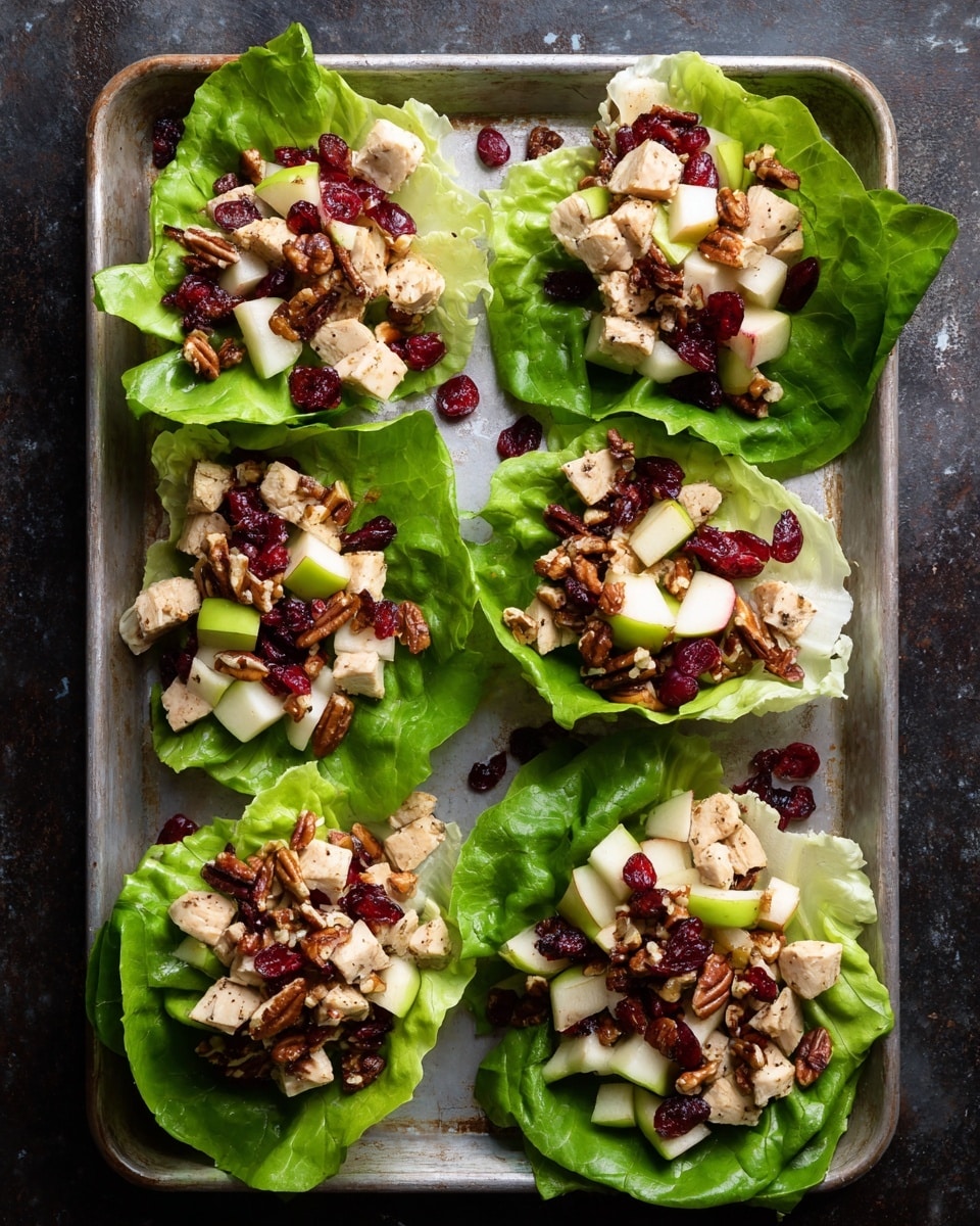 Four lettuce wraps are laid out on a silver baking sheet over a dark textured surface. Each wrap has a bright green, leafy lettuce base with a fresh, crisp texture. On top of each lettuce leaf, there is a chunky mixture of diced light beige chicken, small white apple cubes, red dried cranberries, and brown pecan pieces that add a rough texture. The layers contrast well with the shiny metal tray and dark background. The scene looks fresh and colorful, showing each ingredient clearly. Photo taken with an iphone --ar 4:5 --v 7