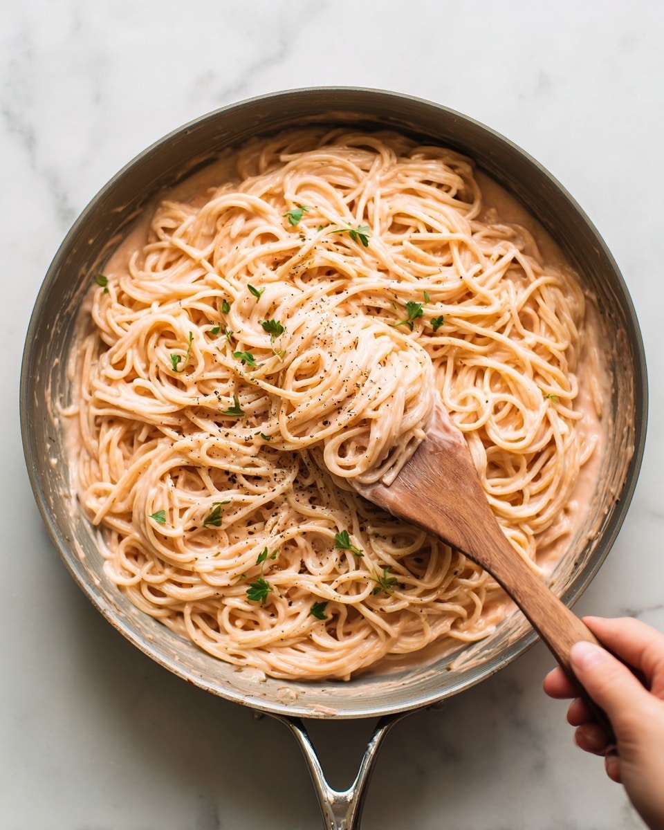 A close-up view of a pan filled with creamy pasta coated in a light pink sauce, with long spaghetti noodles mixed evenly. There are small green herb pieces sprinkled on top along with some black pepper specks. A wooden spoon is lifting some pasta on the right side, and a woman's hand is holding the spoon gently. The pan sits on a white marbled surface. photo taken with an iphone --ar 4:5 --v 7