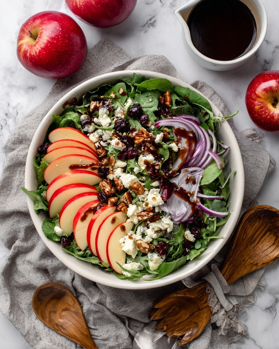 A white bowl filled with a fresh salad, starting with a base layer of mixed green leaves like spinach and arugula. On top, there are bright red apple slices arranged in three areas across the salad, with thin purple onion slices scattered around. Small dark dried berries and chopped brown nuts are sprinkled throughout, along with small white chunks of soft cheese. A dark dressing is drizzled over the salad, adding contrast. The bowl rests on a light gray cloth with a white marbled surface underneath, surrounded by two whole red apples, a white bowl with dark dressing and a spoon, and wooden utensils nearby. photo taken with an iphone --ar 4:5 --v 7