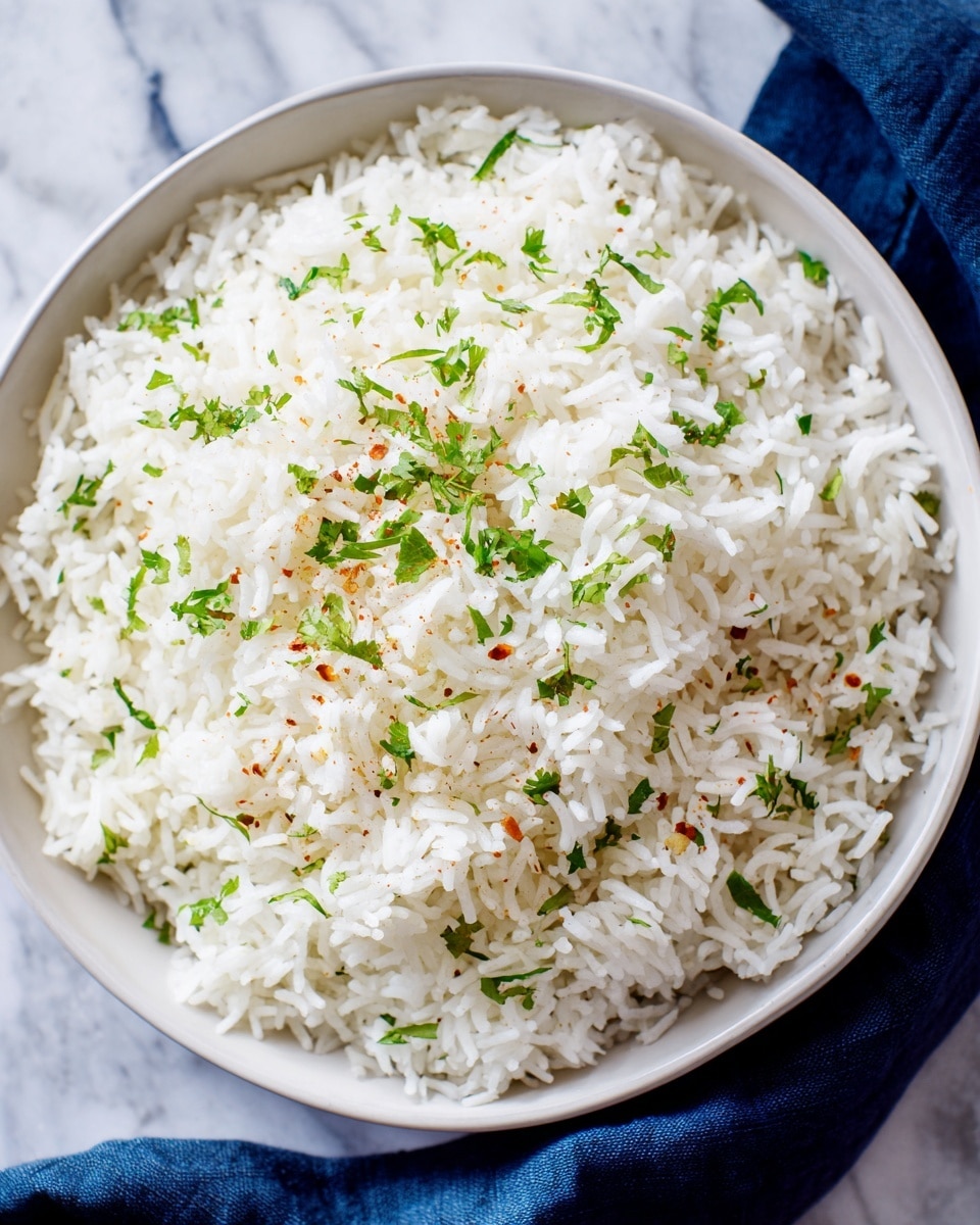 A close-up top view of a bowl filled with white rice, with small green herbs sprinkled evenly on top for color and freshness, along with light reddish-brown spices scattered over the surface. The bowl is white and placed on a white marbled surface, with a dark blue cloth partly visible on the side. The rice grains are fluffy and separated, and the green herbs add texture and brightness. Photo taken with an iphone --ar 4:5 --v 7