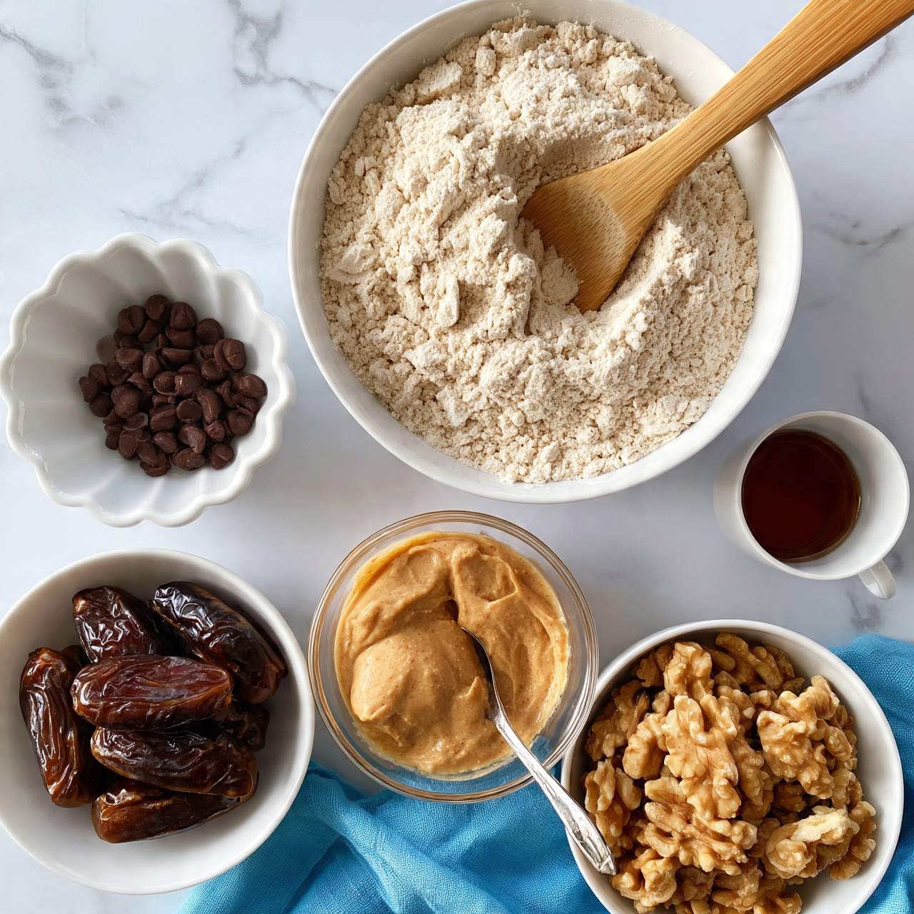 square image The image shows six bowls with different ingredients placed on a white marbled surface. At the top, there is a large white bowl filled with a light beige powdery flour mixture and a wooden spoon resting inside. To the left, a white scalloped bowl contains small dark brown chocolate chips. Below it, a white bowl holds several glossy dark brown dates. To the right of the dates is a clear glass bowl with smooth light brown nut butter and a spoon inside. Next to that, a small white cup has a dark amber liquid, likely syrup. At the bottom right, a white bowl is filled with large light brown walnut halves. A blue cloth is partially visible under some of the bowls. The scene is bright and clean, simple and organized photo taken with an iphone --ar 4:5 --v 7