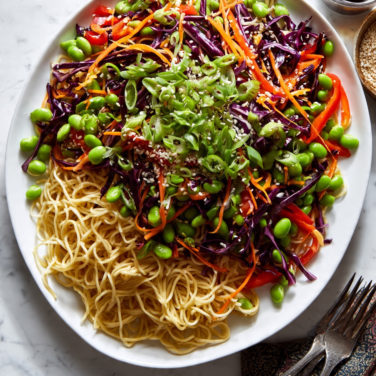 square image A large white oval plate holds a vibrant noodle dish with three main layers visible: the bottom layer is thin, light yellow noodles twisted across the plate; the middle layer is a mix of colorful vegetables including thin strips of orange carrots, lengths of dark purple cabbage, bright green edamame, and slices of red bell pepper scattered evenly; the top layer is garnished with fresh green sliced scallions and a sprinkle of white sesame seeds adding texture. The plate sits on a white marbled surface, with a small portion of a dark patterned cloth and two forks at the bottom right edge of the plate, and a bowl of sesame seeds and a glass container with a dark sauce partially visible in the upper right corner. photo taken with an iphone --ar 4:5 --v 7