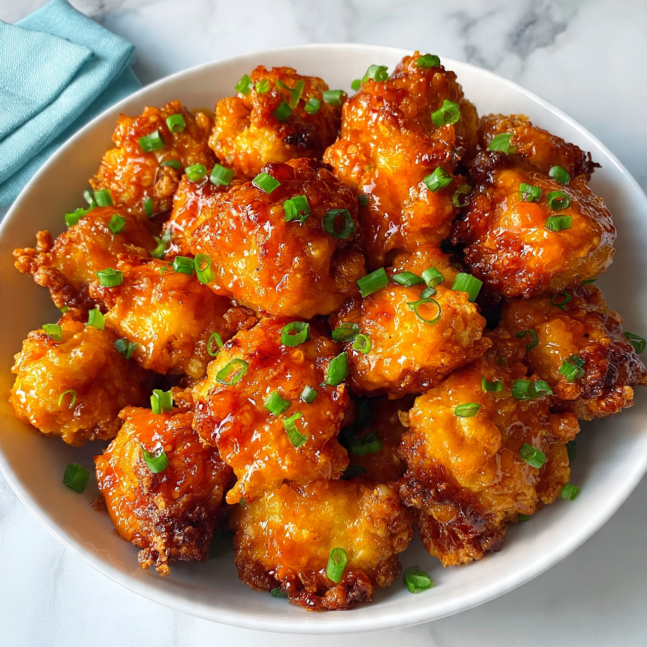 square image The image shows a white bowl filled with many pieces of golden-brown fried chicken bites coated in a shiny, sticky orange sauce. The chicken pieces have a rough, crispy texture and are sprinkled with small green onion slices evenly over the top, adding a fresh green contrast. The bowl sits on a white marbled surface, and there is a folded light blue cloth napkin nearby. The scene is brightly lit, highlighting the glossy sauce and crispy edges of the chicken. Photo taken with an iphone --ar 4:5 --v 7